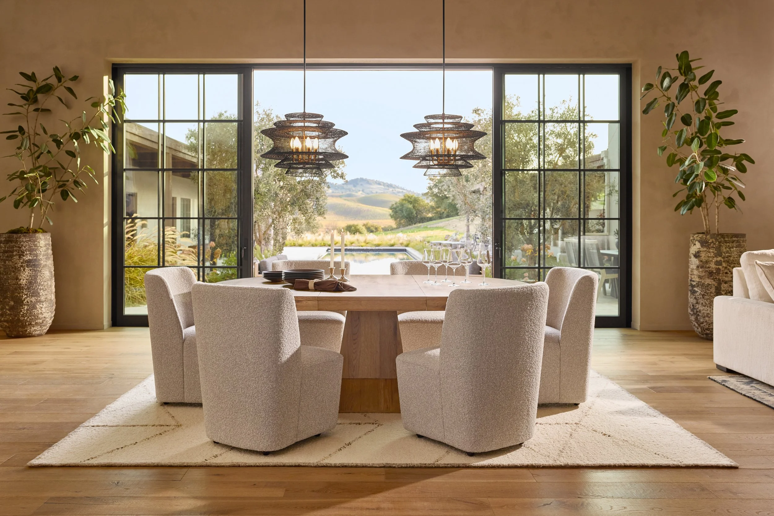 A dining room with a wooden table set with black plates, wine glasses, and a candelabrum, surrounded by six cream-colored upholstered chairs, with large windows showing an outdoor landscape with trees, hills, and a swimming pool.