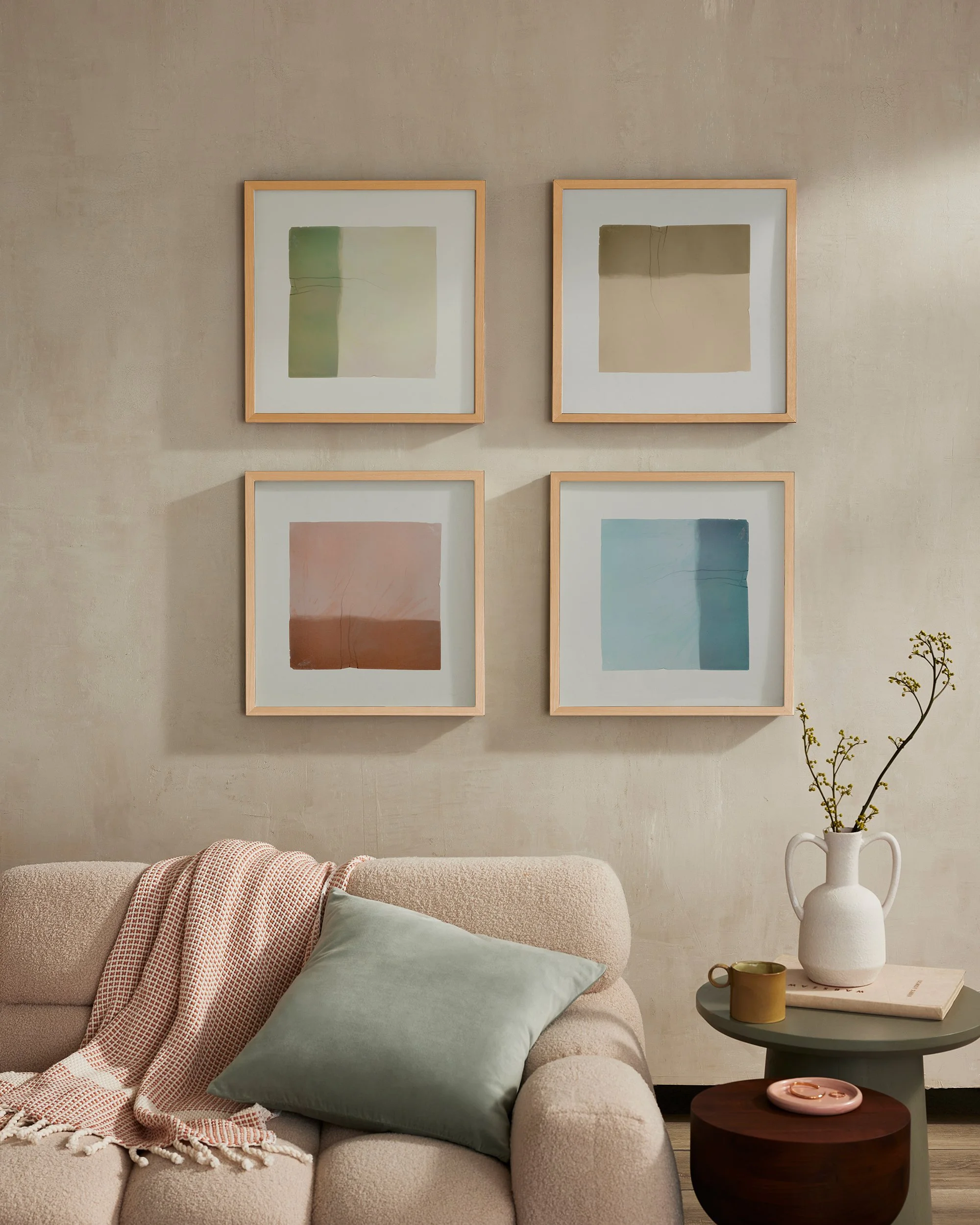 Contemporary living room with a neutral-colored sofa adorned with a pastel green cushion and a pink and white throw blanket. Two square framed abstract art pieces with soft, muted colors hang on a textured beige wall above the sofa. A small, round si