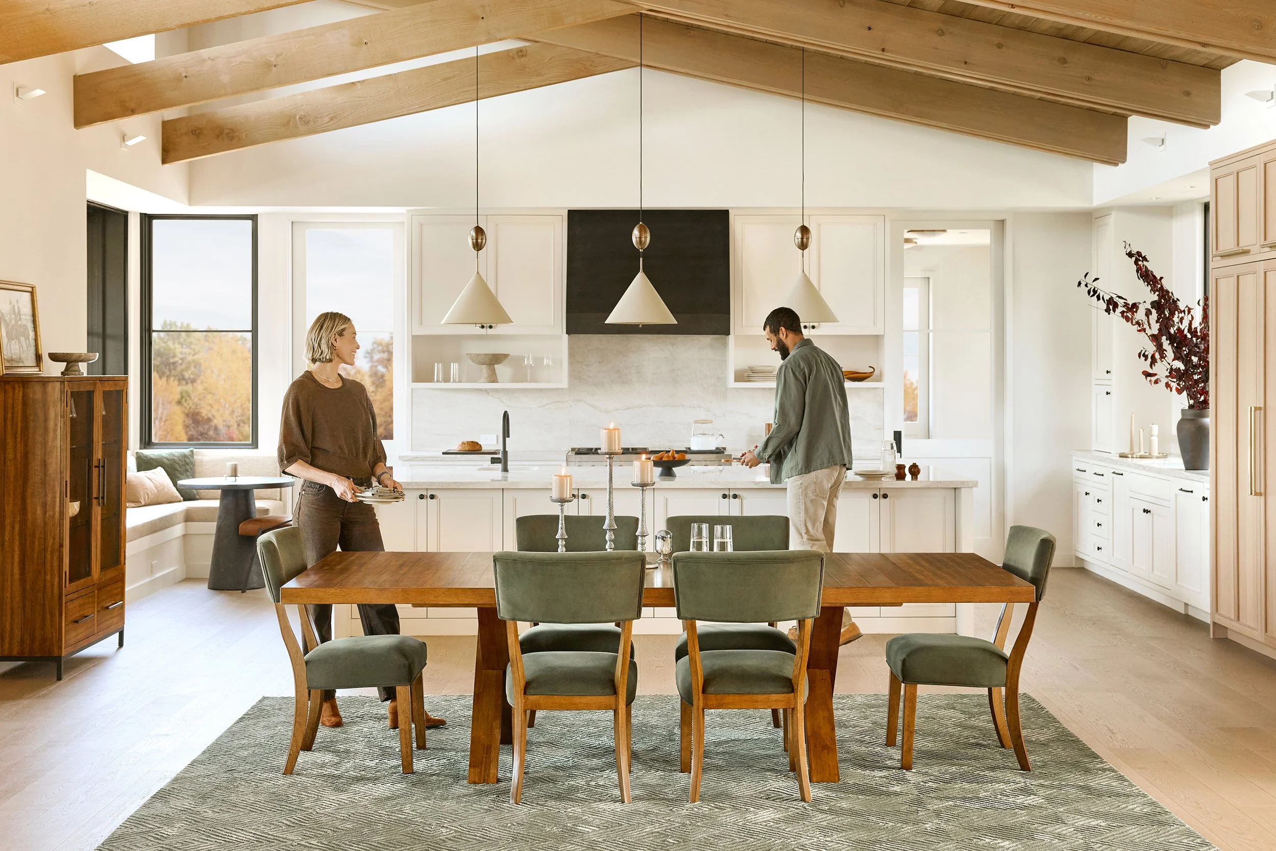 A modern kitchen with a vaulted ceiling and exposed wooden beams. Two people are standing and talking near the kitchen island, which has candles and a coffee cup on it. There are large windows letting in natural light, and a dining table with green c