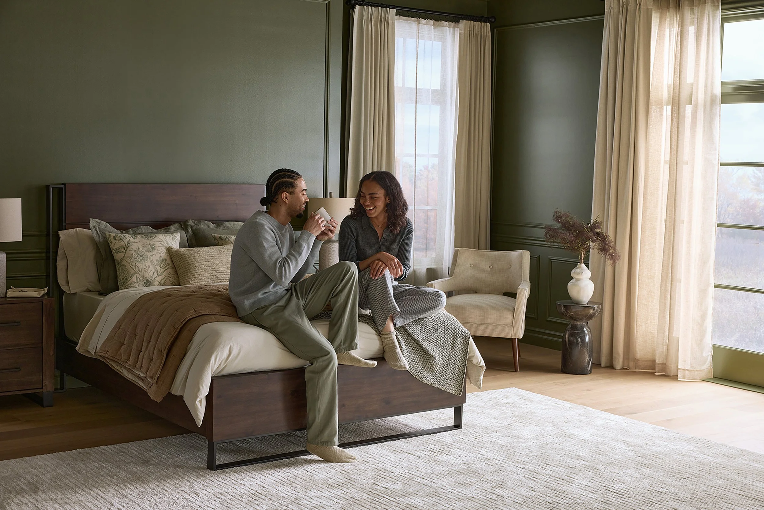 A man and a woman sitting on a bed in a hotel room, smiling and enjoying each other's company, with sunlight streaming through large windows with cream curtains.