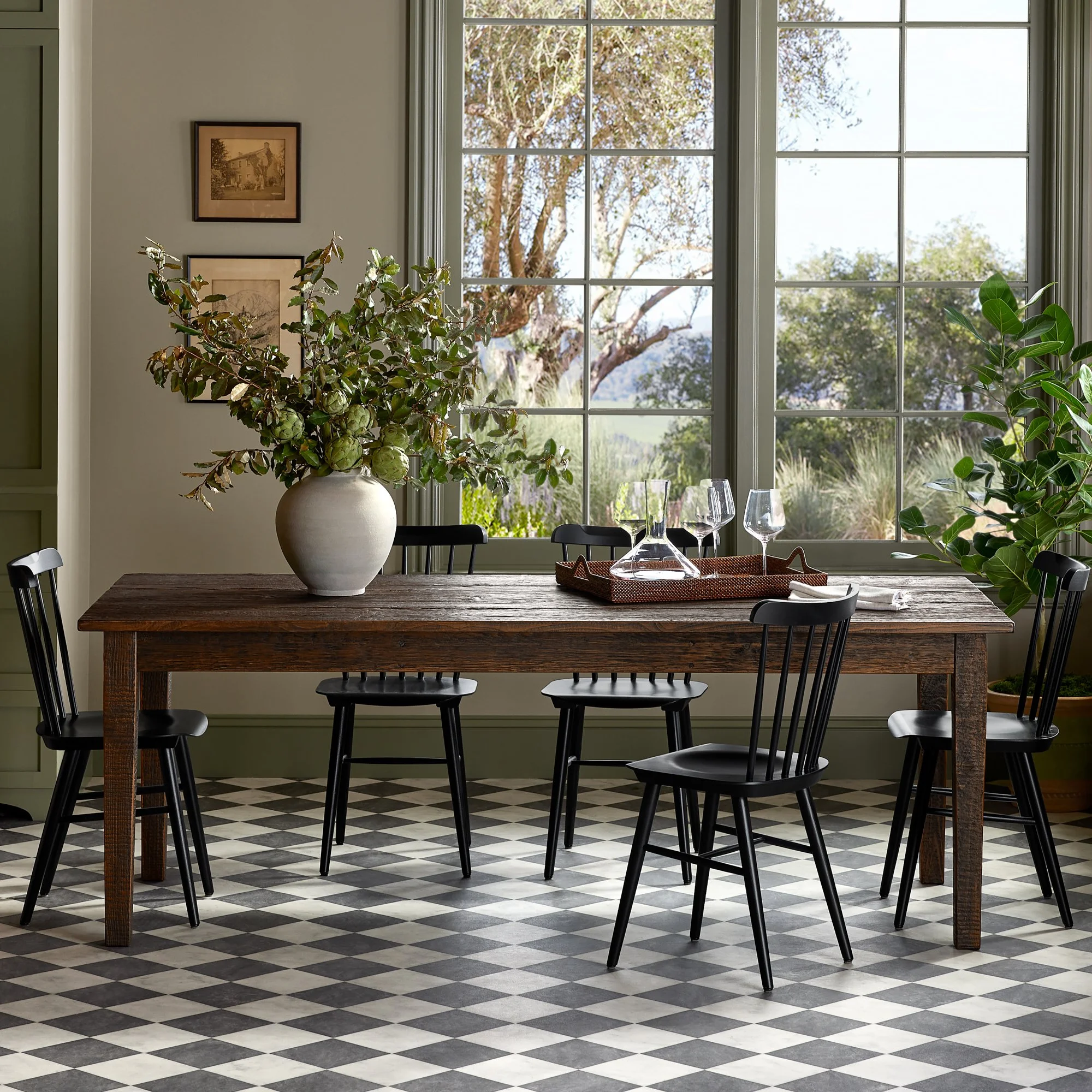 A dining room with a large wooden table, black chairs, a white vase with green and brown foliage, a tray with glasses, and a large window showing trees outside.