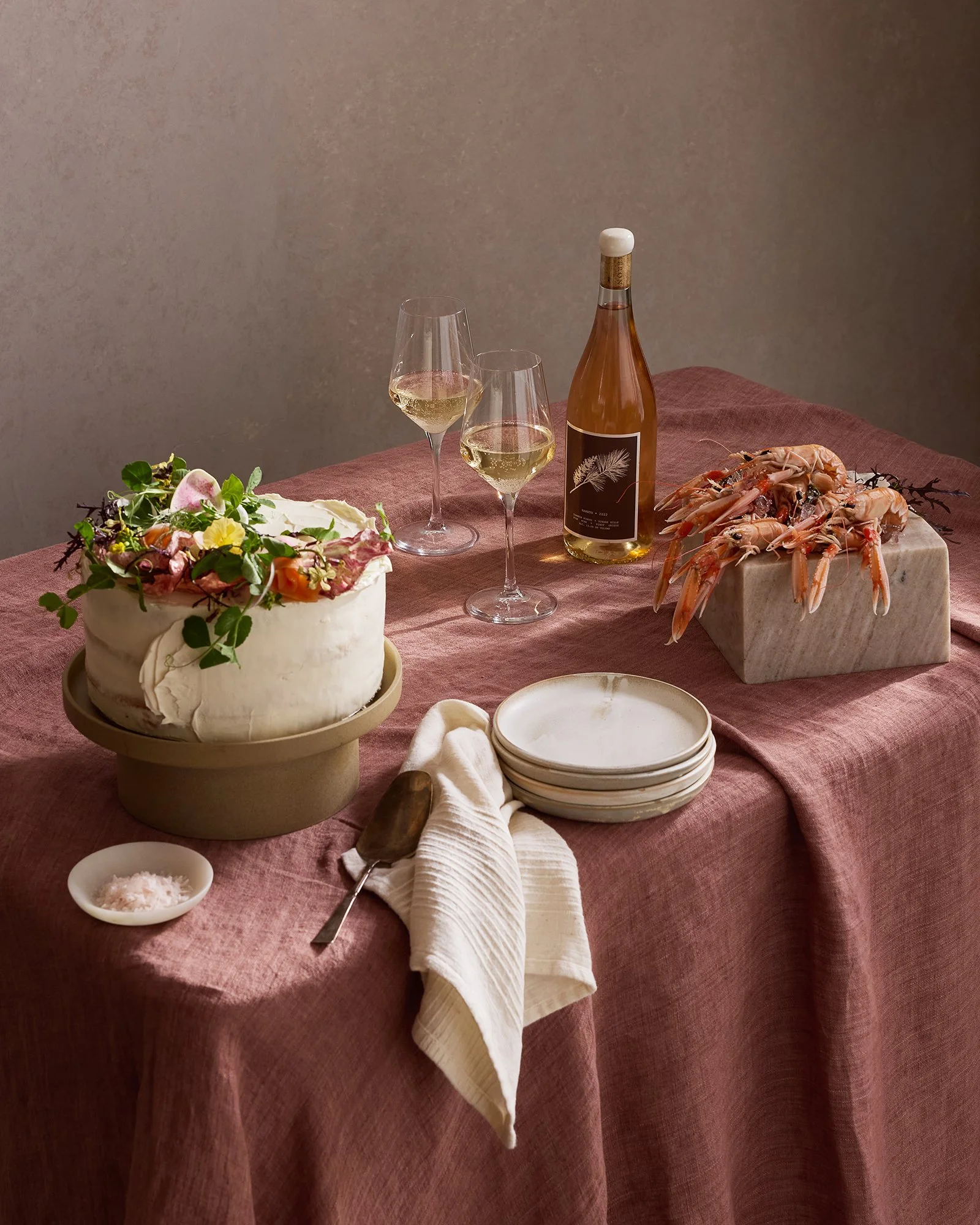 A rustic dining table set with a cake topped with flowers, two glasses of white wine, a bottle of rosé wine, a bowl of pink salt, a serving of shrimp on a marble slab, and a small stack of plates with a linen napkin and a wooden serving utensil.