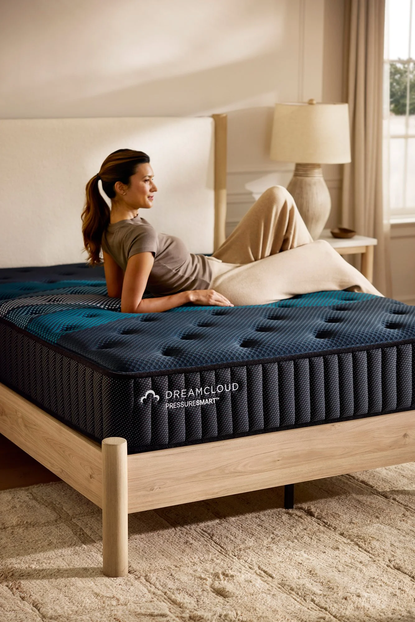 A woman relaxing on a bed with a black and blue mattress, in a well-lit bedroom with beige walls, a beige headboard, a round beige table lamp, and a window with beige curtains.