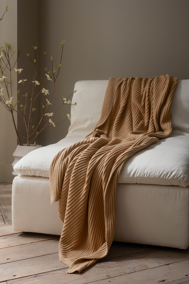 A beige sofa with a brown, ribbed blanket draped over it, located in a room with wooden floors and a tall potted plant with white flowers in the background.