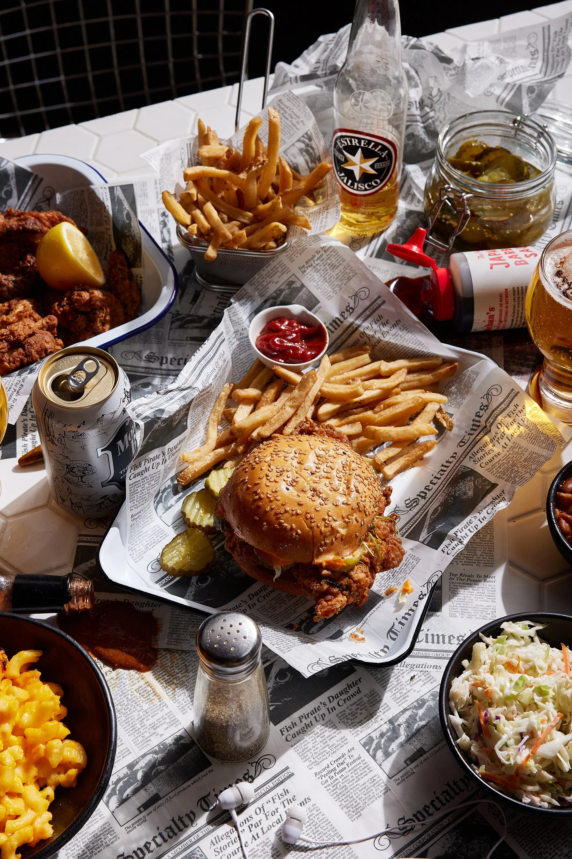 A table with various foods including a fried chicken sandwich with pickles, French fries with ketchup, a bowl of macaroni and cheese, coleslaw, a small bowl of ketchup, a can of beer, a bottle of beer, and a jar of pickles, all on newspaper-style pap