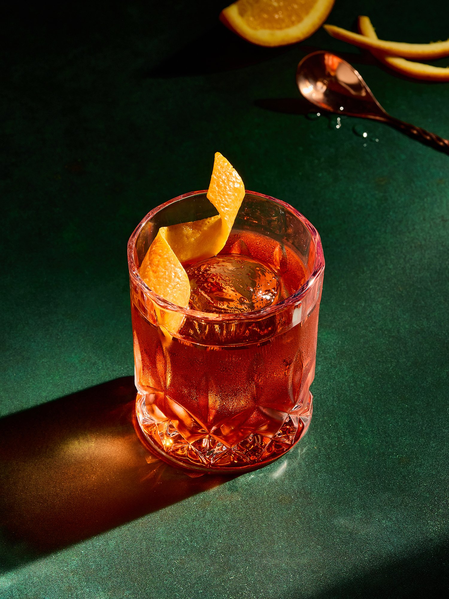 A pink glass with a faceted design contains a drink with an orange slice garnish, placed on a dark green surface. A lemon slice and a peel are partially visible in the background, along with a spoon.