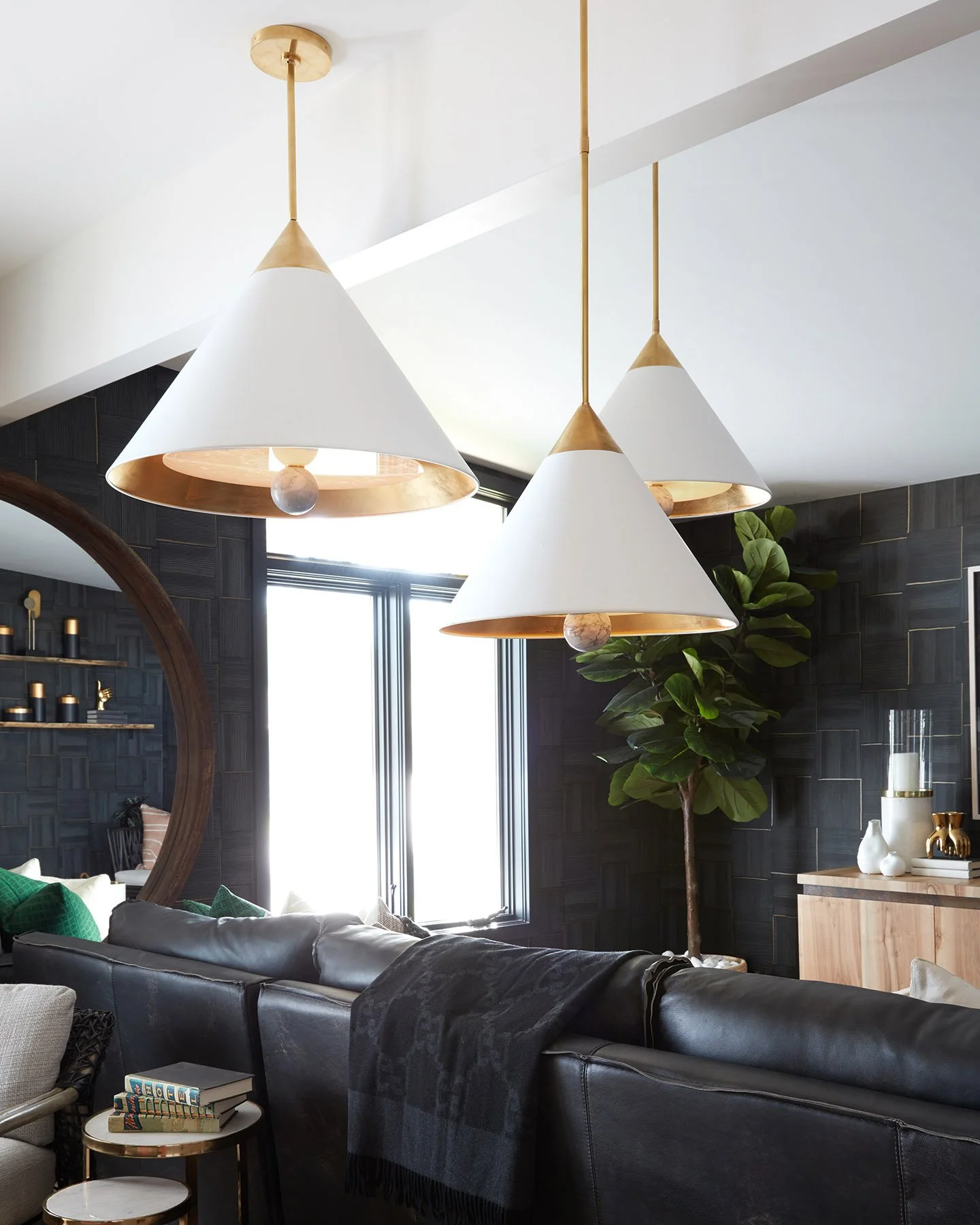 Modern living room with three white conical pendant lights with gold accents hanging from the ceiling, a black leather sofa, a small side table with books, a large window, a tall green plant, and dark textured wall panels.