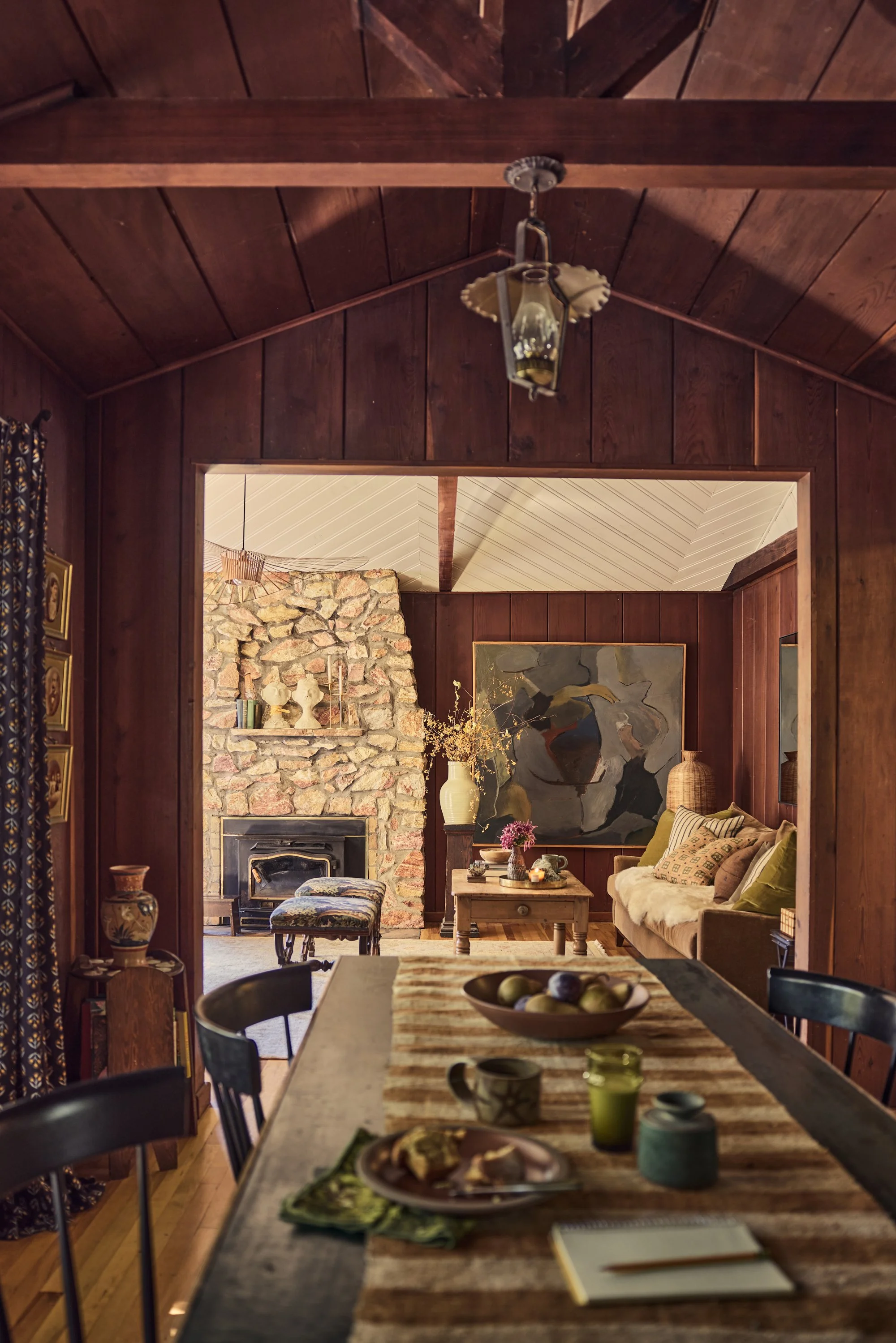 Cozy living room with stone fireplace, dark wood walls, and a ceiling with exposed beams. Features a painting of a bird, a lamp, and decorative items on shelves.