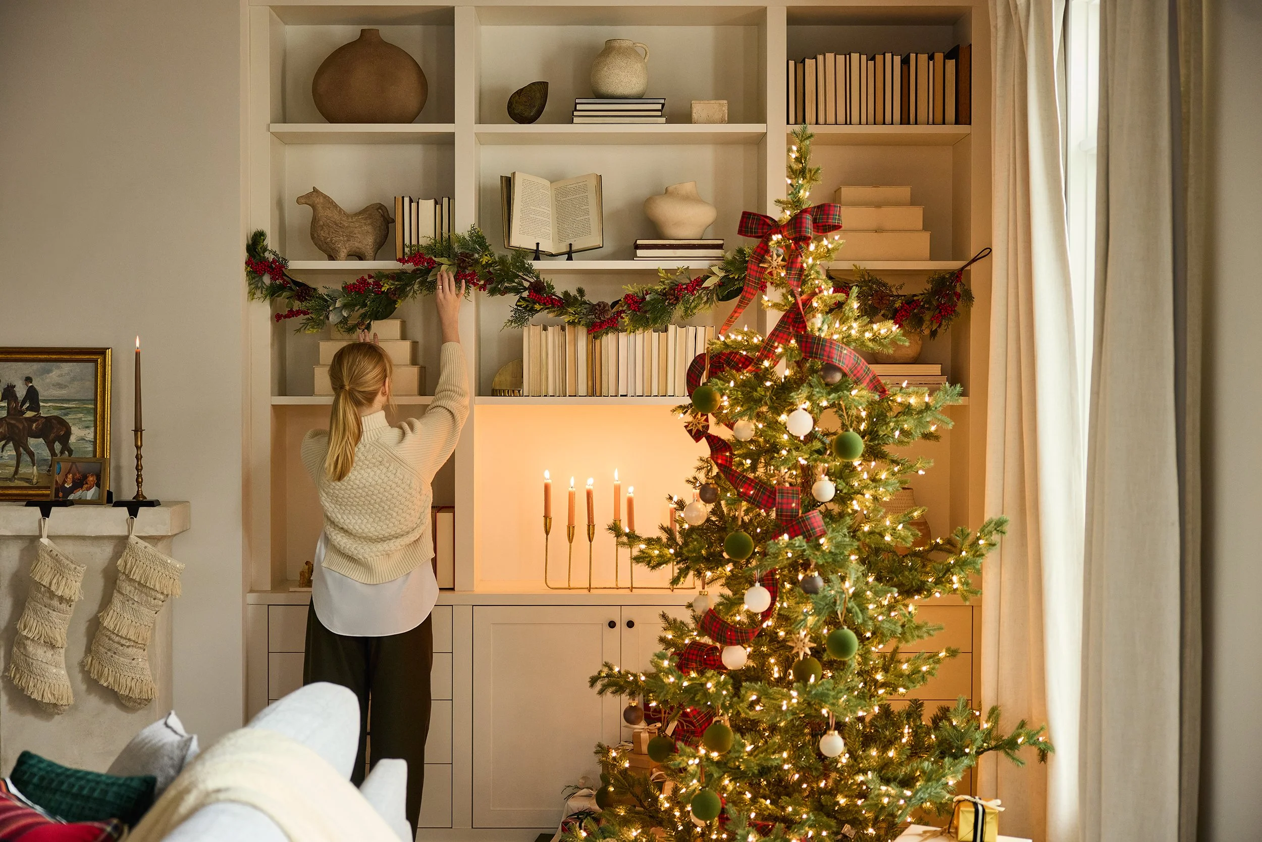 A woman decorating a Christmas mantel with greenery and red berries, with a Christmas tree to the right decorated with plaid ribbons and white and green ornaments, and a fireplace with lit candles in front.
