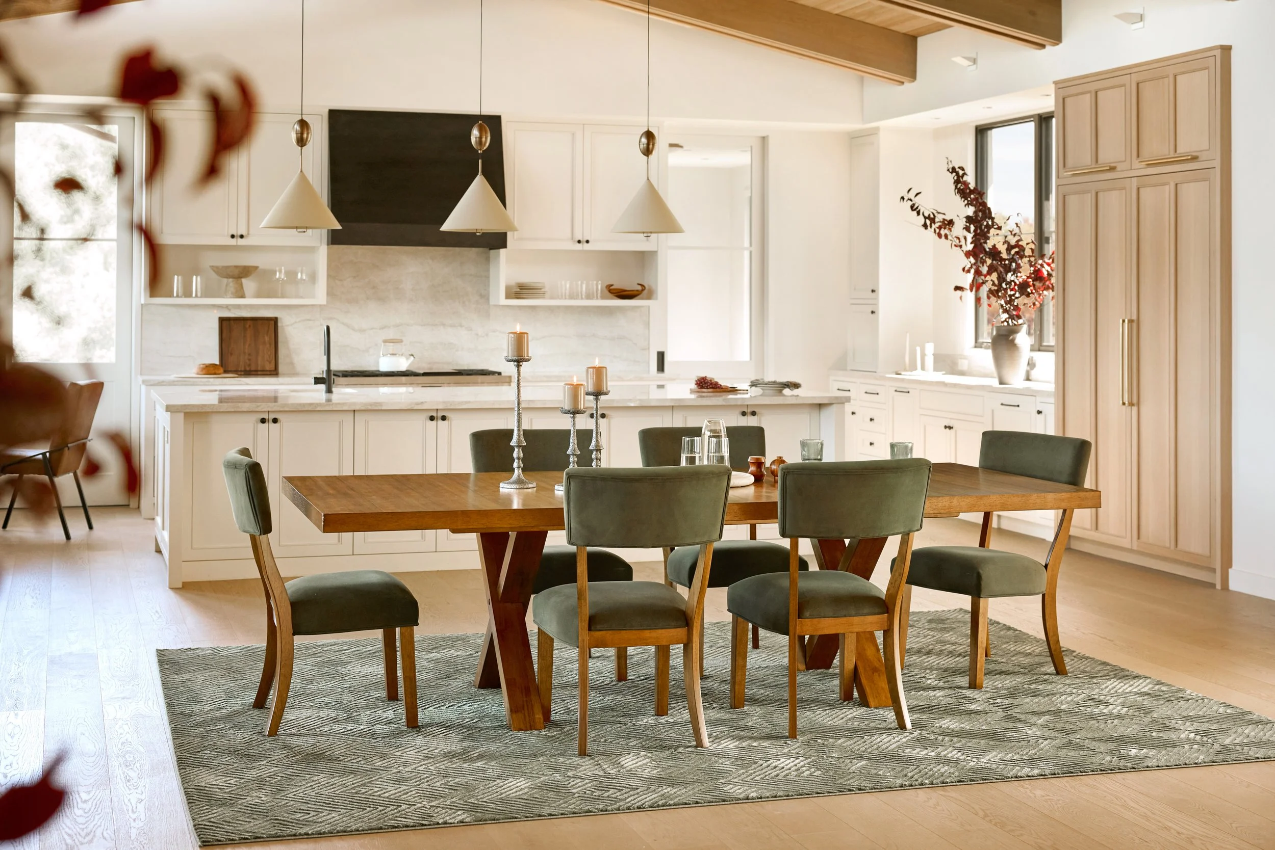 A dining room with a wooden table surrounded by six green upholstered chairs, a gray patterned rug underneath, and candles and glassware on the table. In the background, an open kitchen with white cabinets, a marble countertop, a black oven hood, and