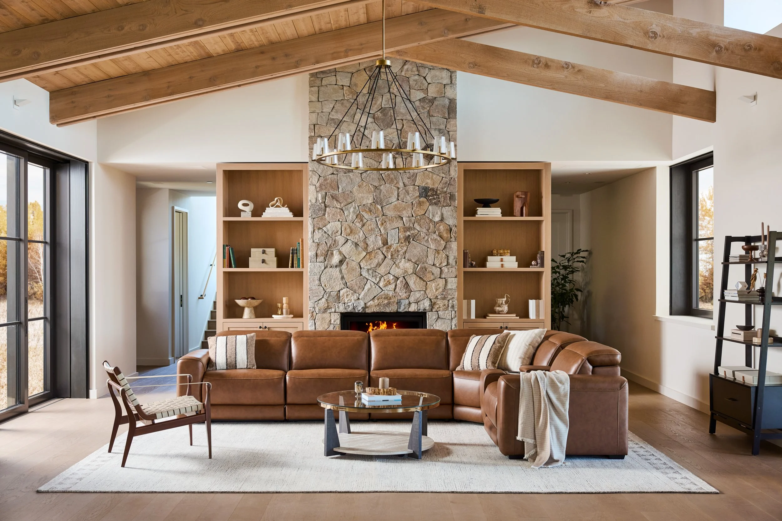 Living room with a stone fireplace, wooden ceiling beams, large windows, a brown leather sofa, a wooden armchair, a round glass coffee table, and built-in bookshelves.