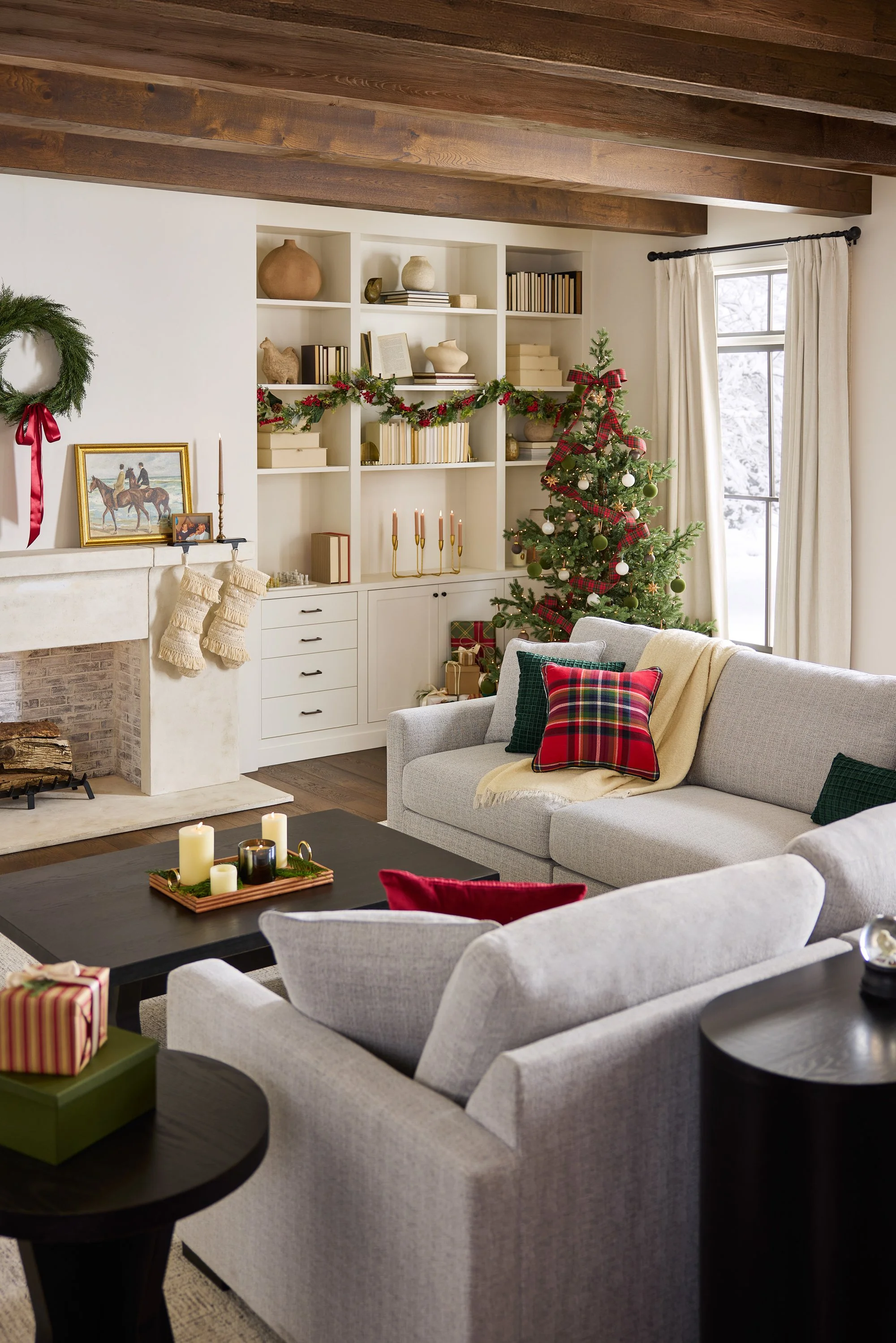 A cozy living room decorated for Christmas with a small decorated Christmas tree, a white built-in bookshelf with garland, and a fireplace with stockings, a framed picture, and candles on the mantle. There are white and gray sofas with holiday pillow