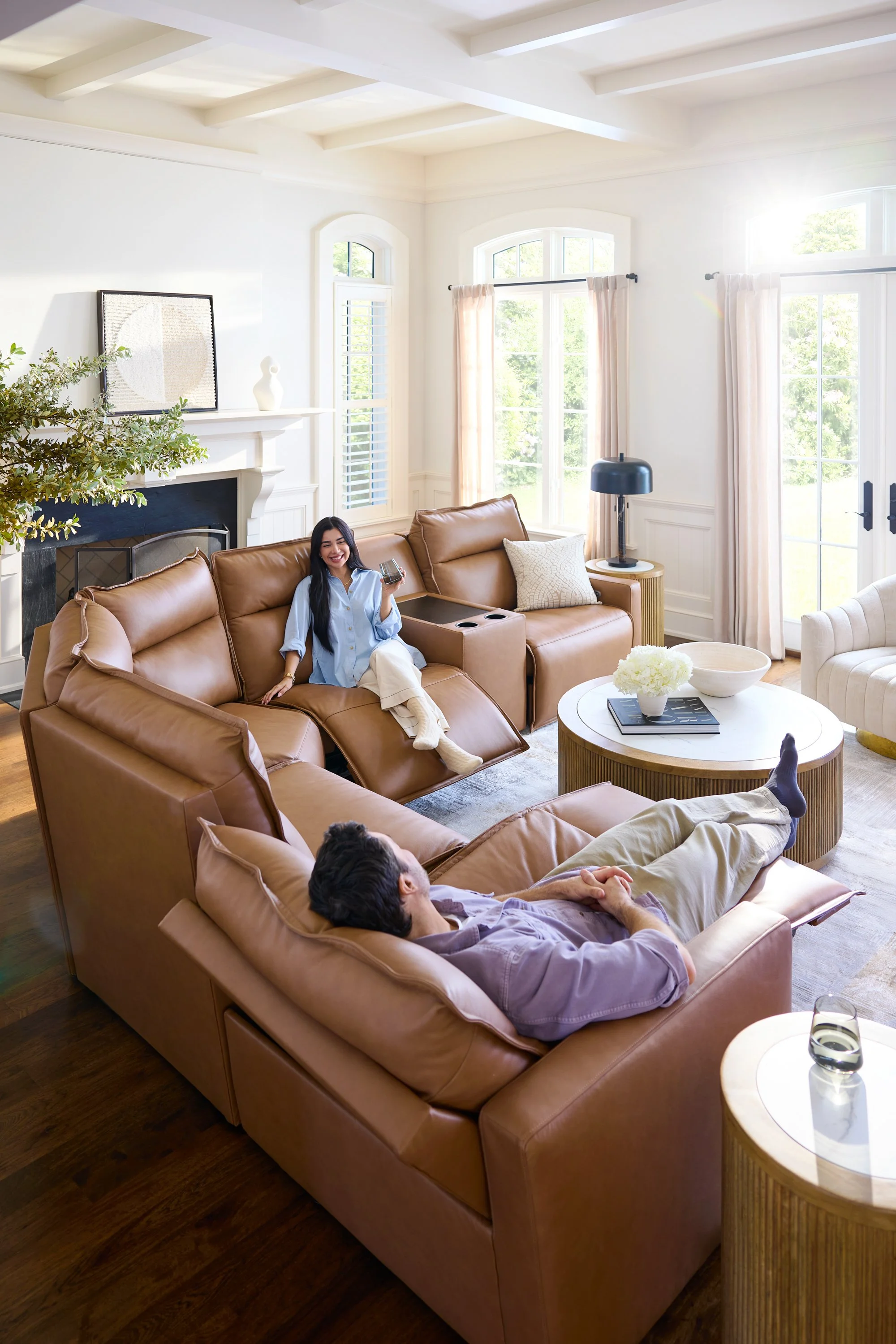 Living room with large windows, beige sectional sofa with two people sitting, one woman smiling with a drink, one man lying down, coffee table with flowers, and a fireplace.