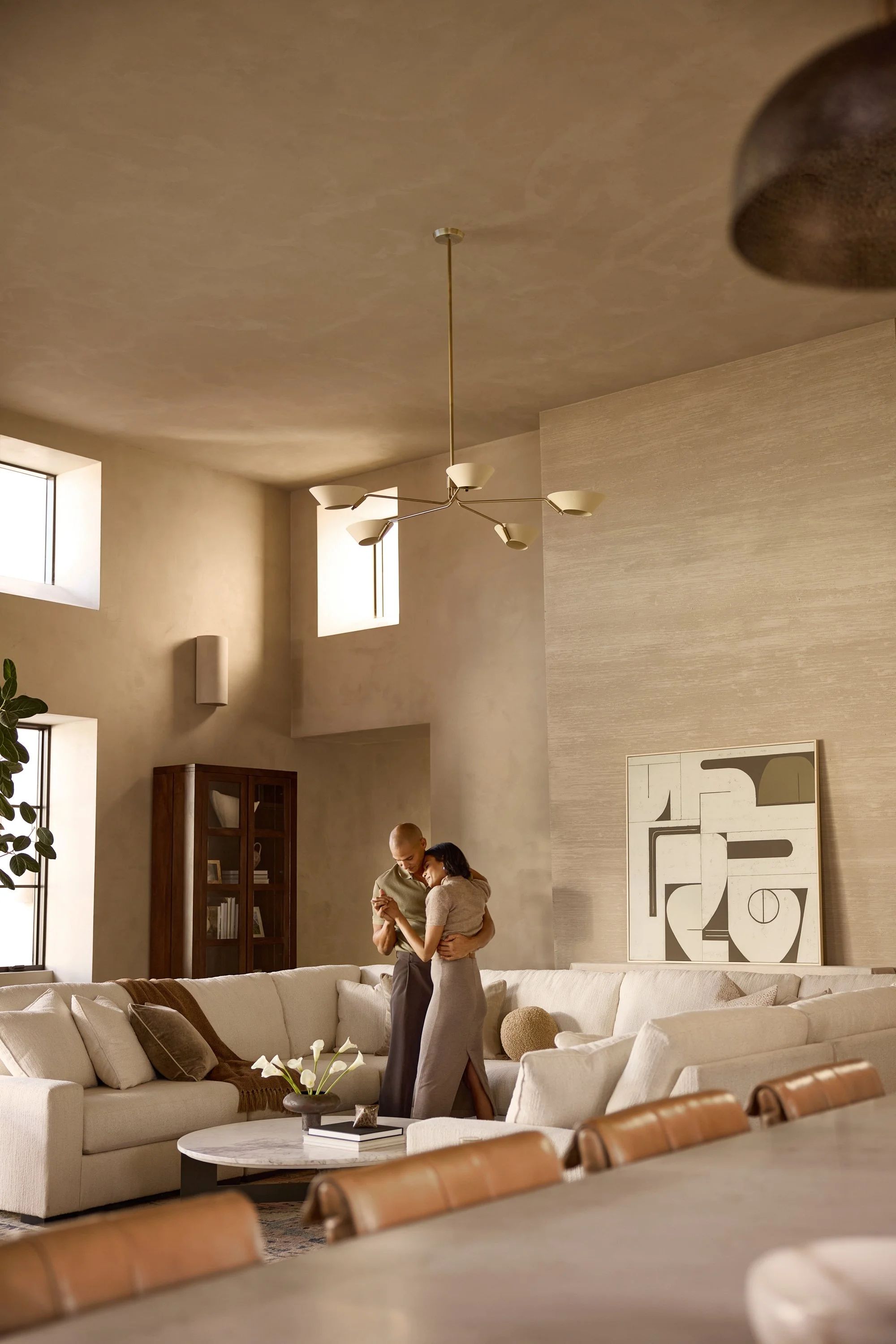 A couple hugging and looking at a smartphone in a modern living room with a beige sectional sofa, abstract wall art, wooden cabinet, and large windows.