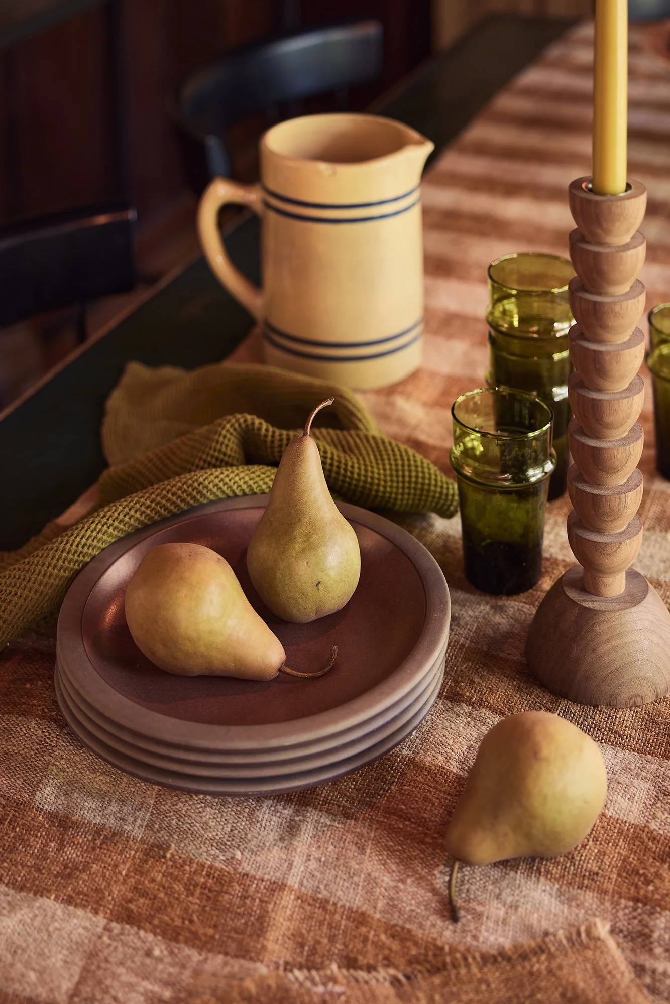 A table with a ceramic pitcher, green glasses, pears, a yellow candle, and a plate with a cloth on a woven tablecloth, in a cozy setting.