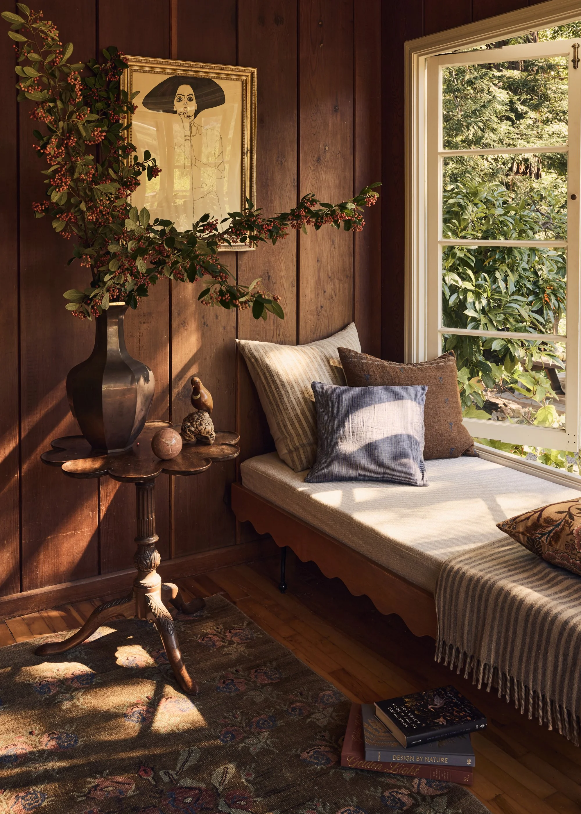 A cozy corner with a wooden wall, a window with an outdoor view of green trees, a cushioned window seat with pillows, a decorative vase with branches and berries, a small table with pebbles and a figurine, a framed artwork of a woman, and a stack of 