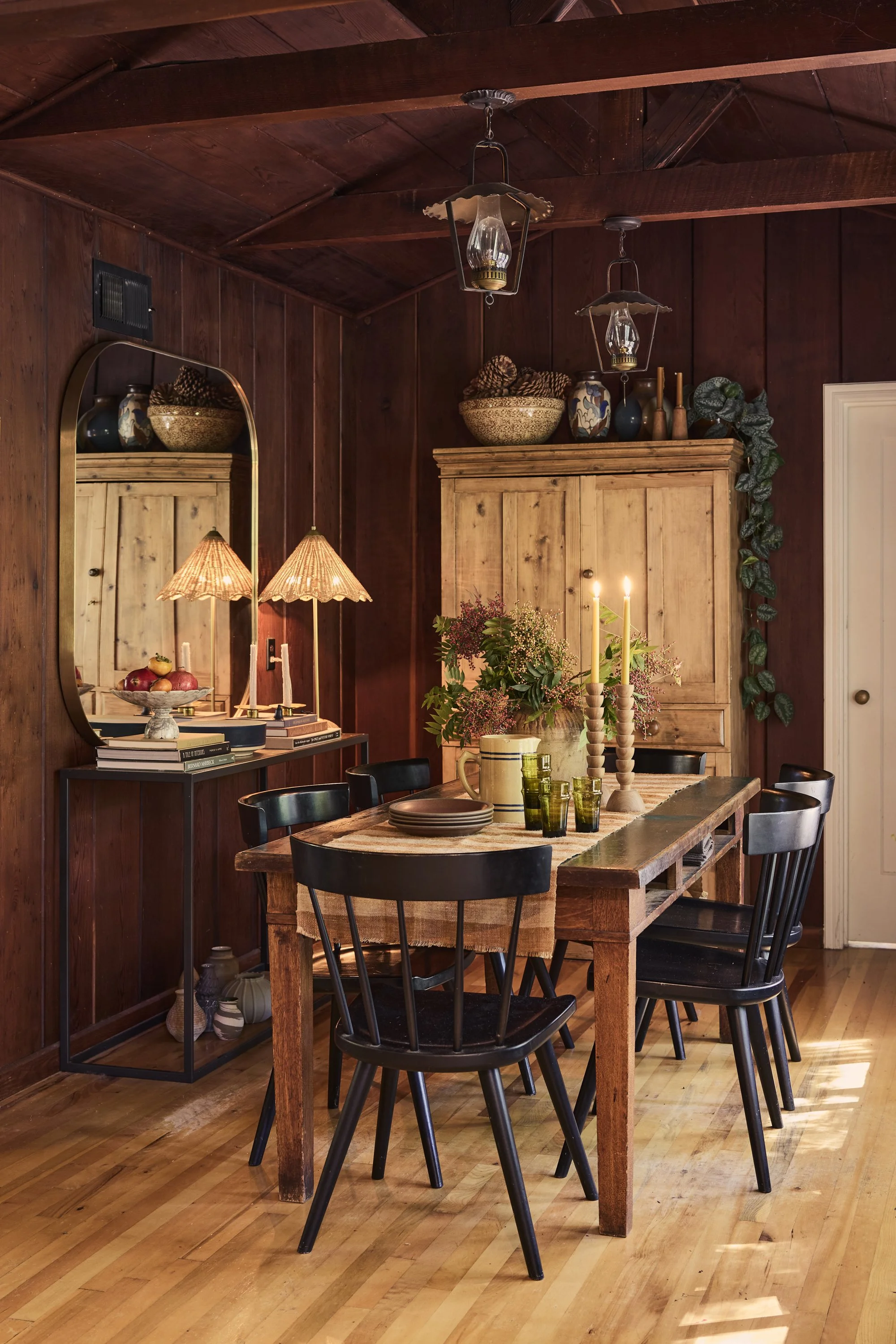 A rustic dining room with a wooden dining table set with green glasses, a cream-colored pitcher, and a floral centerpiece with candles. Black chairs surround the table, and a large wooden cabinet with decorative vases and bowls is against the dark wo