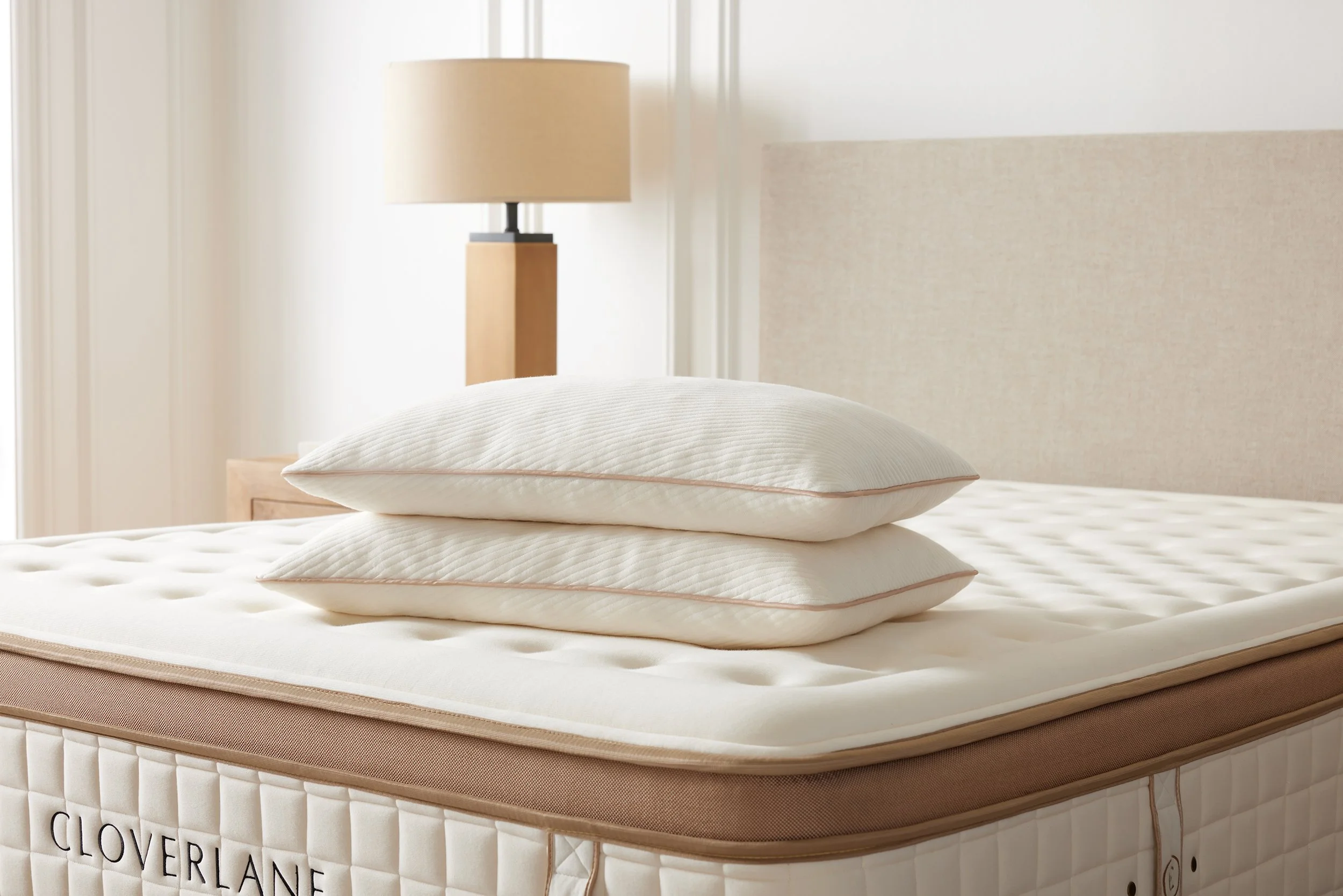 A bed with two stacked pillows, a beige headboard, and a beige table lamp with a black base in a bright room.