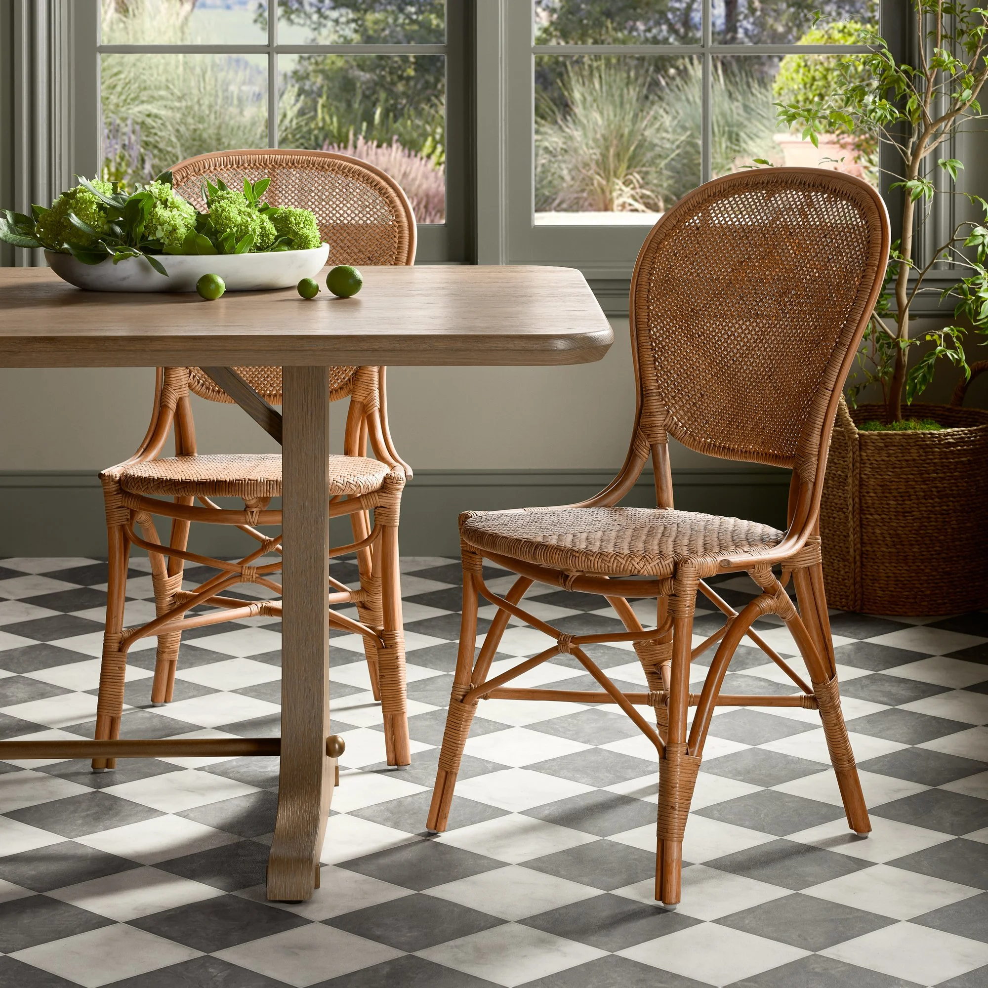 A wooden dining table with a white bowl of green hydrangea flowers and three green limes, set in front of large windows with a garden view, surrounded by wicker chairs and a tall potted plant.