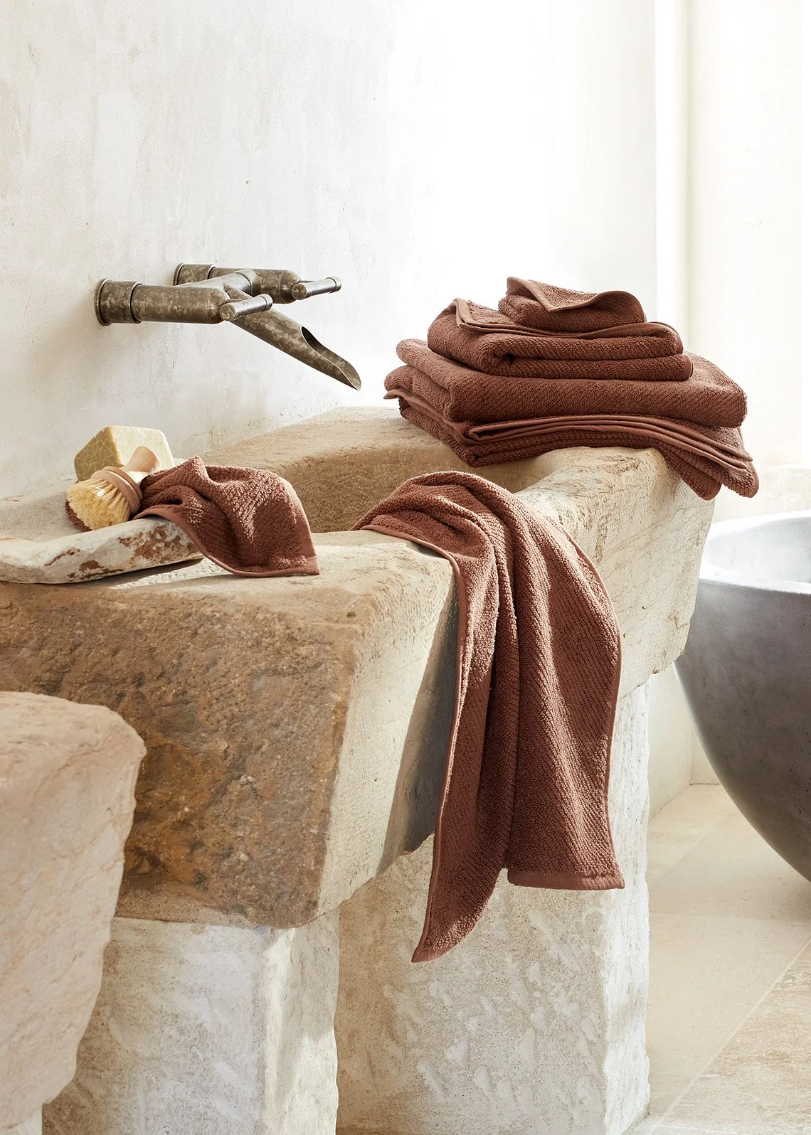 A rustic stone bathtub with folded brown towels on the side and a pile of matching towels on top. A natural sponge and a brush are placed on the edge of the tub. The background is a simple white wall with soft natural light.