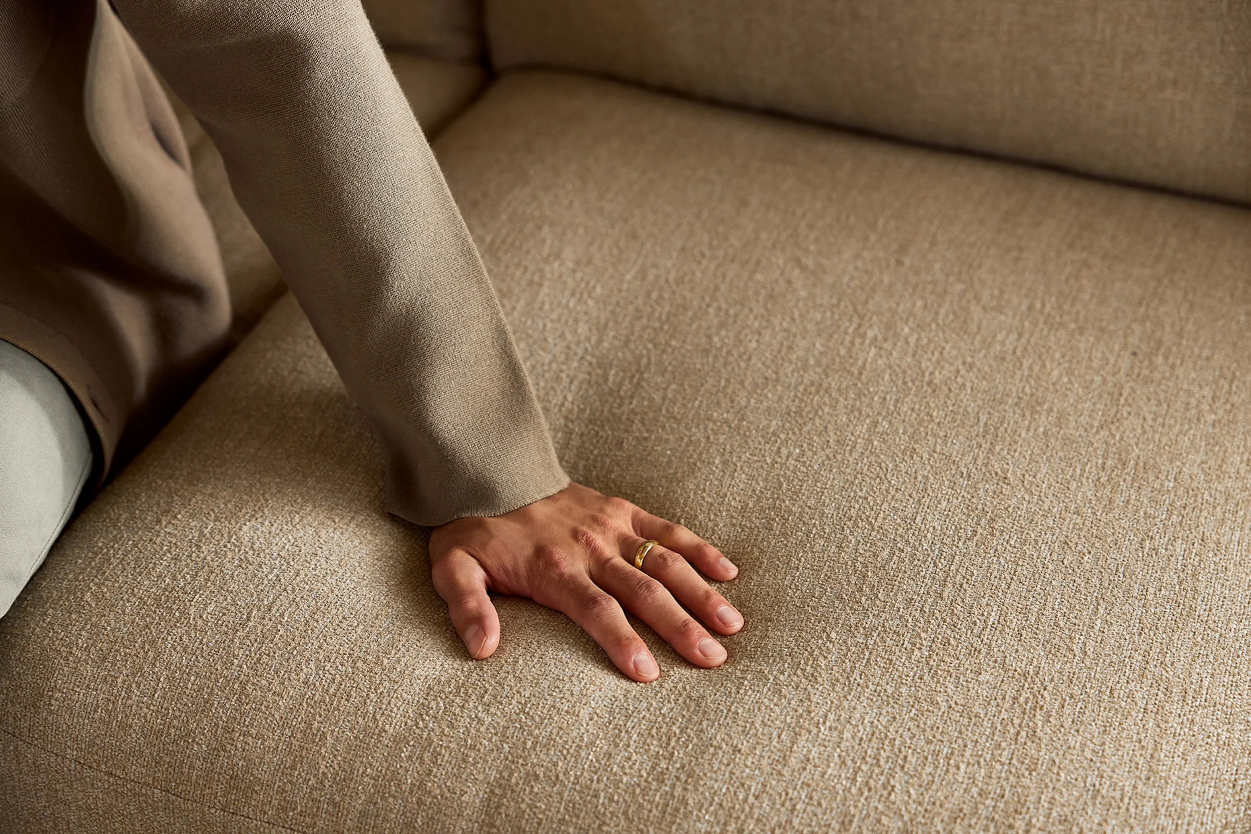 A person is kneeling on a beige textured carpet, with one hand flat on the carpet. The person is wearing a beige or light brown sweater and has a gold ring on the ring finger of their right hand.