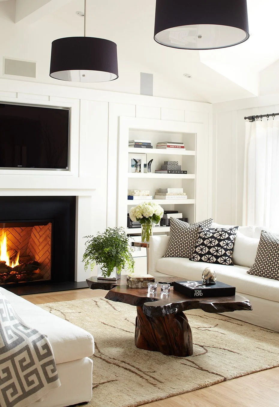 Living room with white sofa, patterned throw pillows, a wooden coffee table, fireplace, flat-screen TV, bookshelf with books, black pendant lights, and large window with sheer curtains.