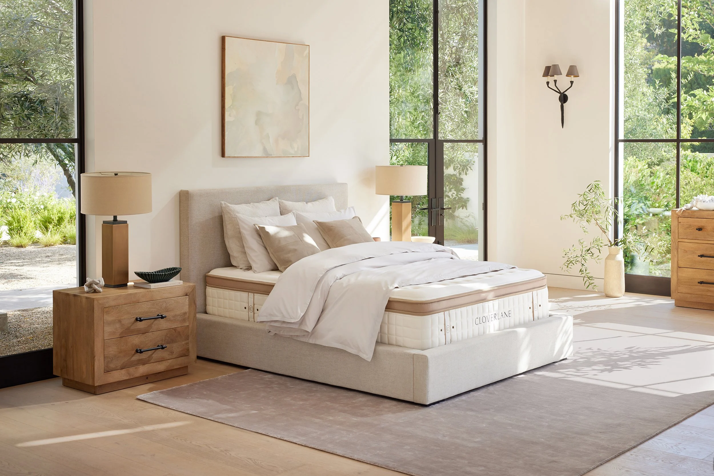 A modern bedroom with large windows showing greenery outside, a bed with cream-colored bedding, wooden nightstands with lamps, a wall art piece, and a wooden dresser.