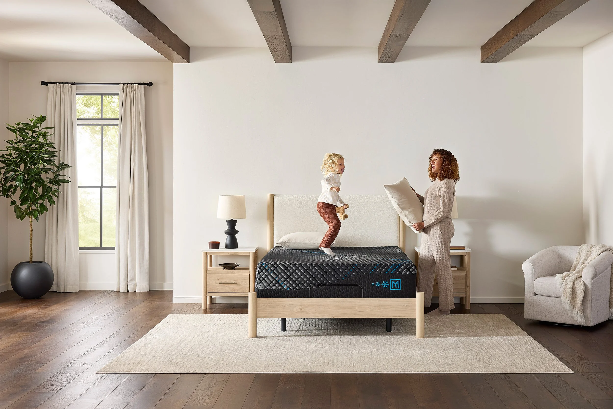 A woman and a young girl are playing on a bed in a bright, minimalist bedroom with white walls, wooden ceiling beams, a large window with curtains, a potted plant, and modern furniture.