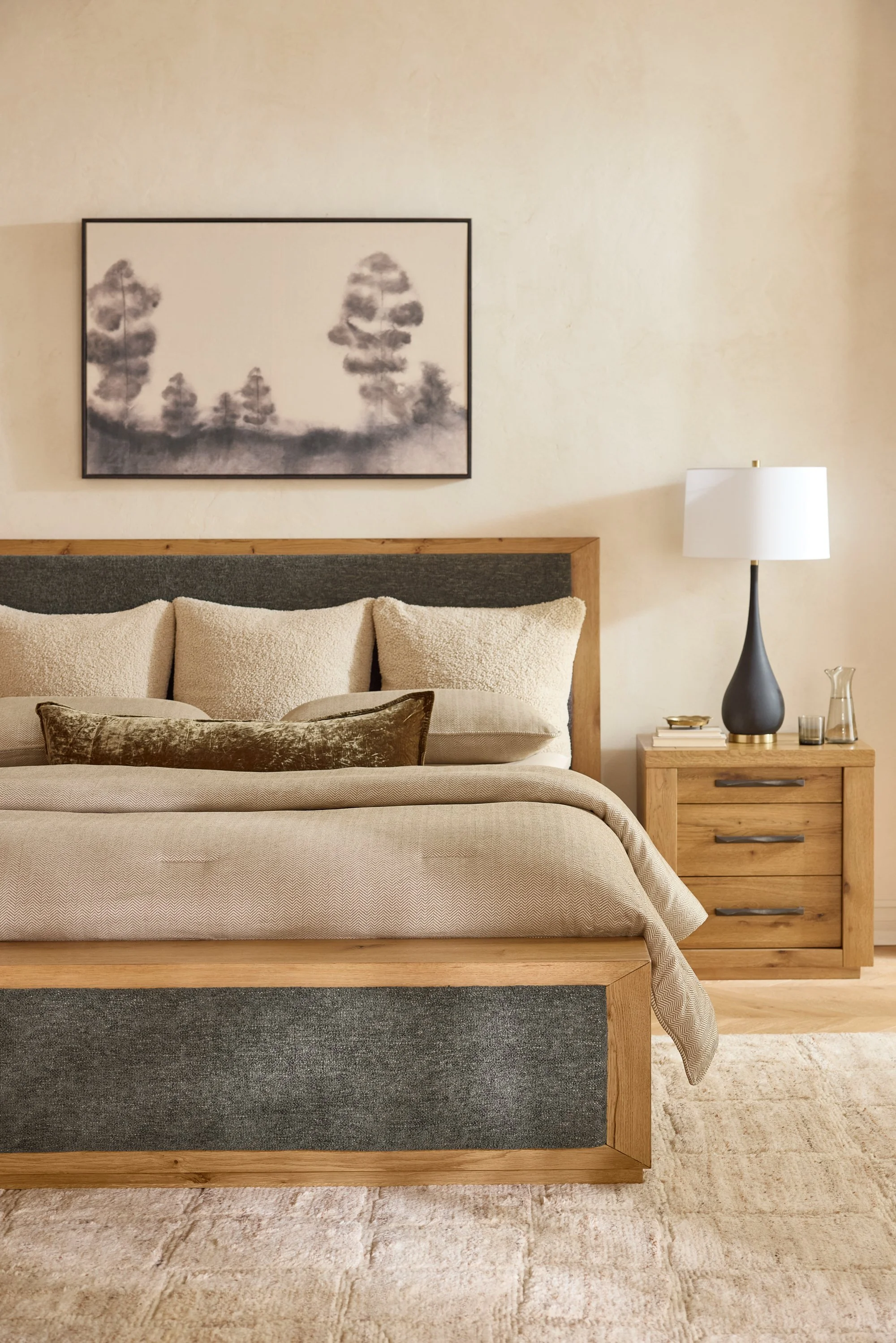 A bedroom with a wooden bed frame, beige bedding, and plush pillows. A black and white abstract landscape painting hangs above the bed. To the right, a wooden nightstand with a black vase-shaped lamp, a small tray, a glass, and some books. A light-co