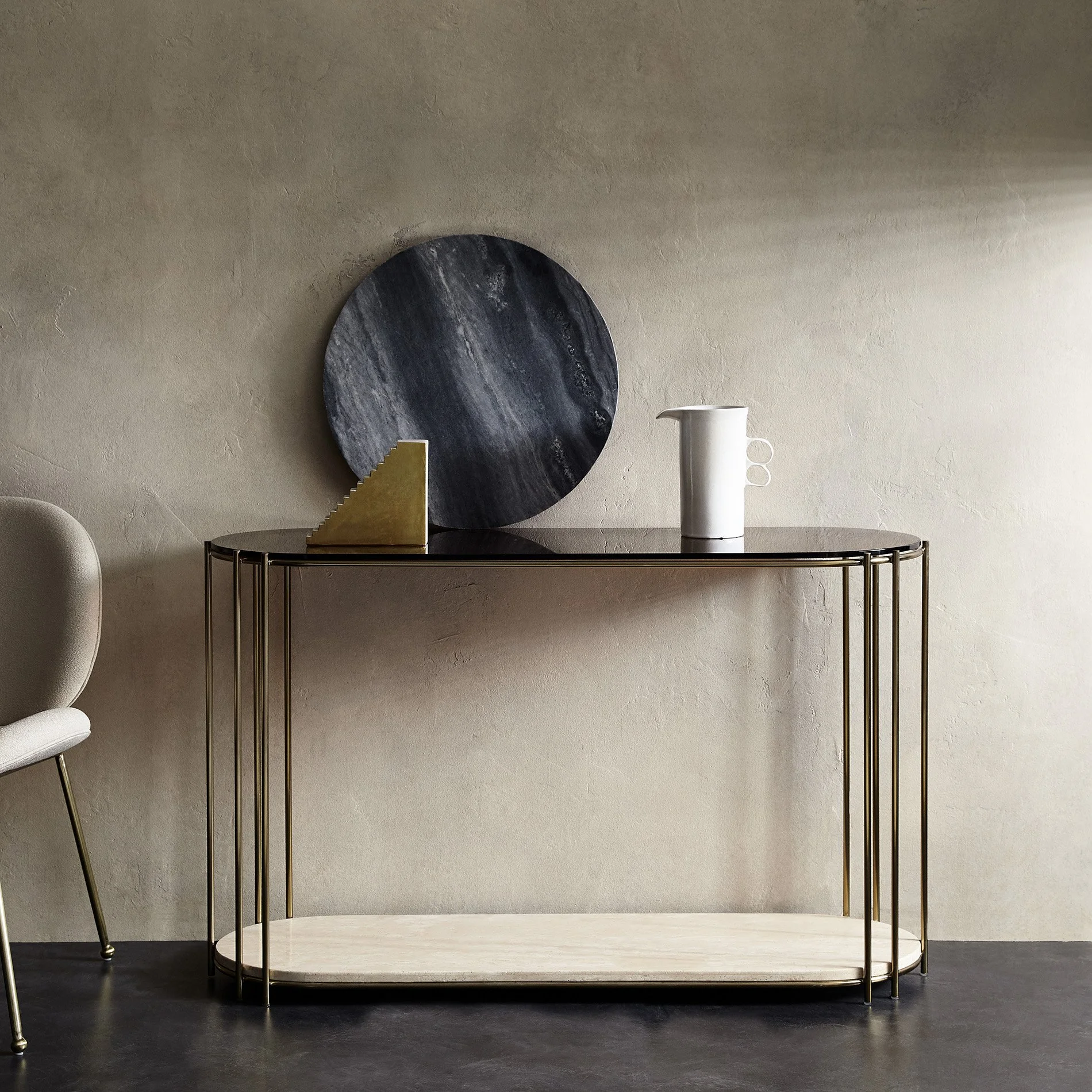 Empty round marble and gold bedside table with a large round art piece on the wall behind it and a white ceramic pitcher with handle and spout on top of the table. The setting is minimalist with a beige wall and dark flooring.
