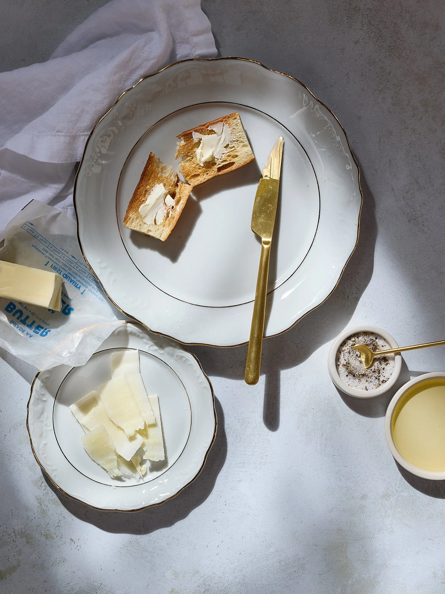 A porcelain plate with a gold rim holding two pieces of toasted bread with butter. There are slices of cheese on a small plate, a packet of butter, a small bowl of salt, and a small bowl of pepper on a light-colored surface with a white cloth in the 