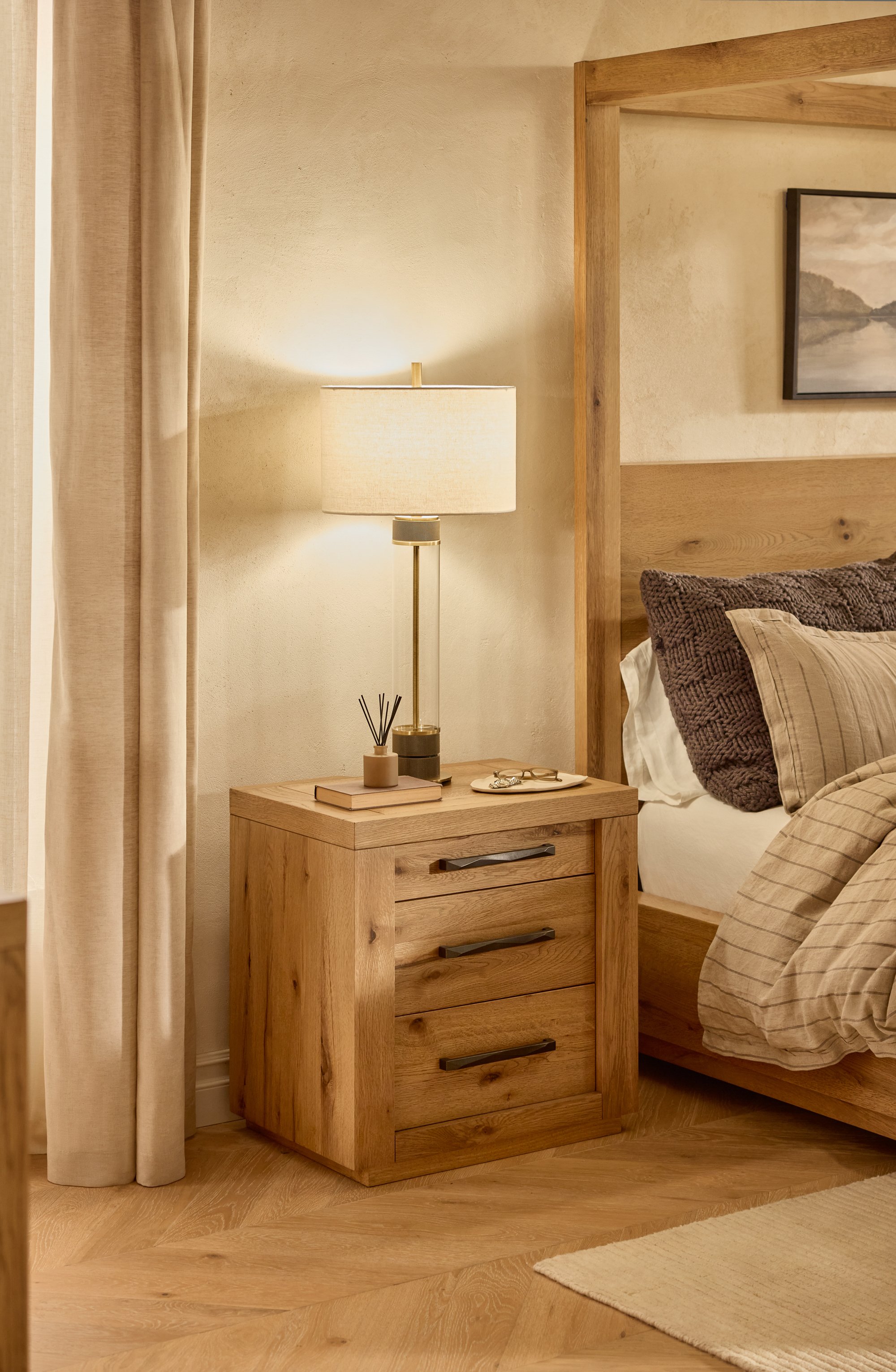 A bedroom with beige walls, a wooden nightstand, a table lamp, and part of a bed with brown pillows and beige linens. There is a framed landscape picture on the wall, beige curtains, and a light-colored rug on the wooden floor.