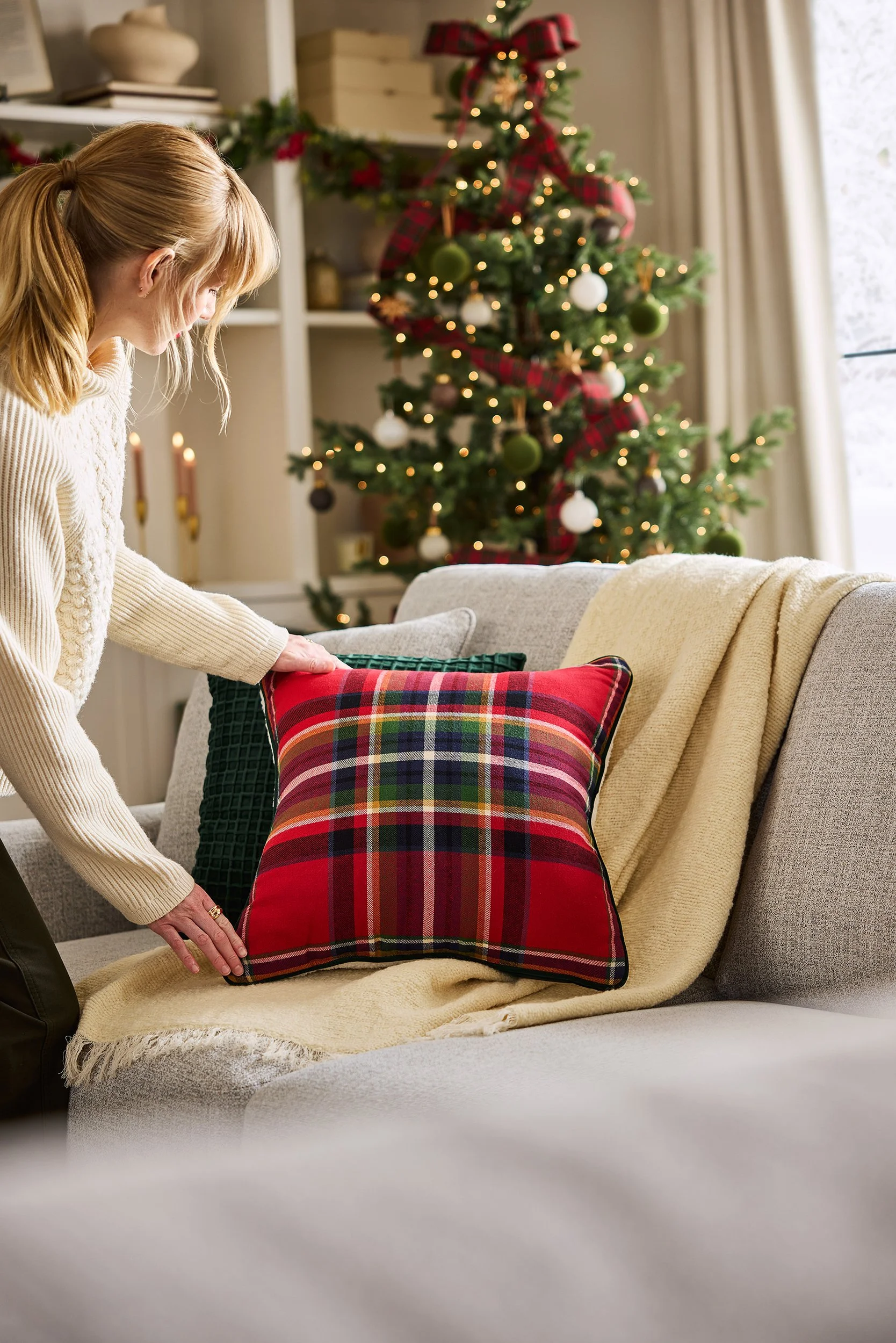 A woman arranging a red plaid Christmas pillow on a sofa with a cream-colored blanket draped over it, in front of a decorated Christmas tree with lights and ornaments, in a cozy living room.