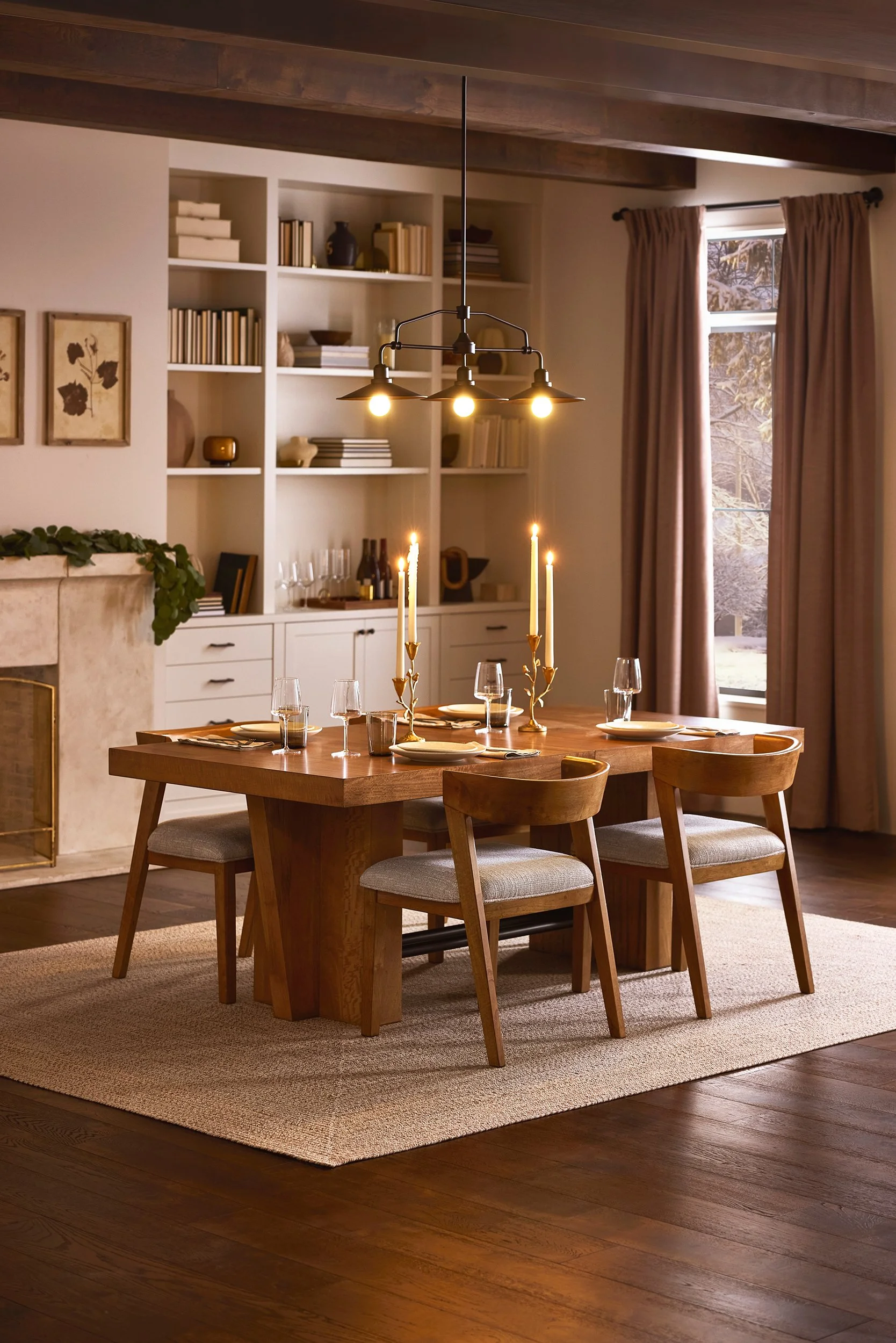 A dining room with a wooden table set for a meal, featuring a candelabra and wine glasses. There is a window with brown curtains, and a built-in white bookshelf filled with books and decorative items. A chandelier with five lights hangs over the tabl