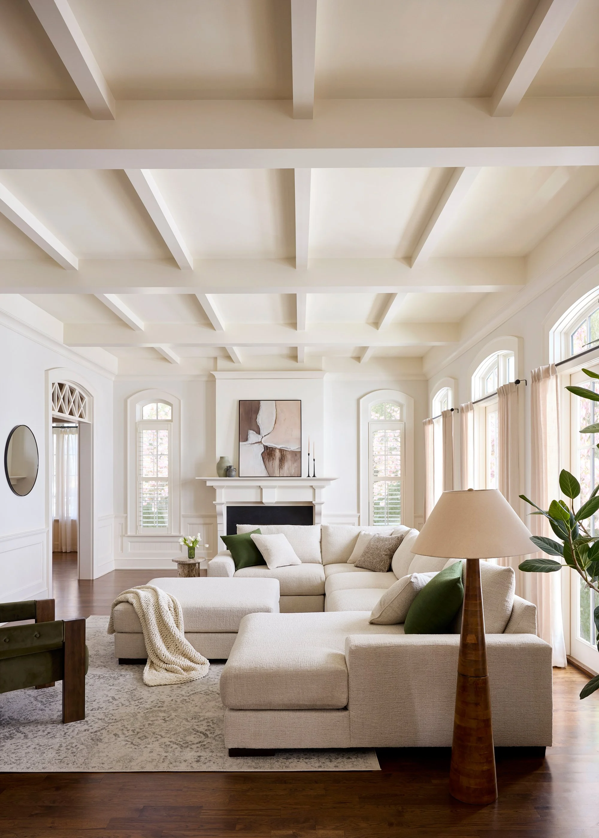 Living room with white walls, high ceilings with white beams, large windows with curtains, a beige sofa with green pillows, a side table with a lamp, a fireplace with abstract artwork above it, and hardwood floors.