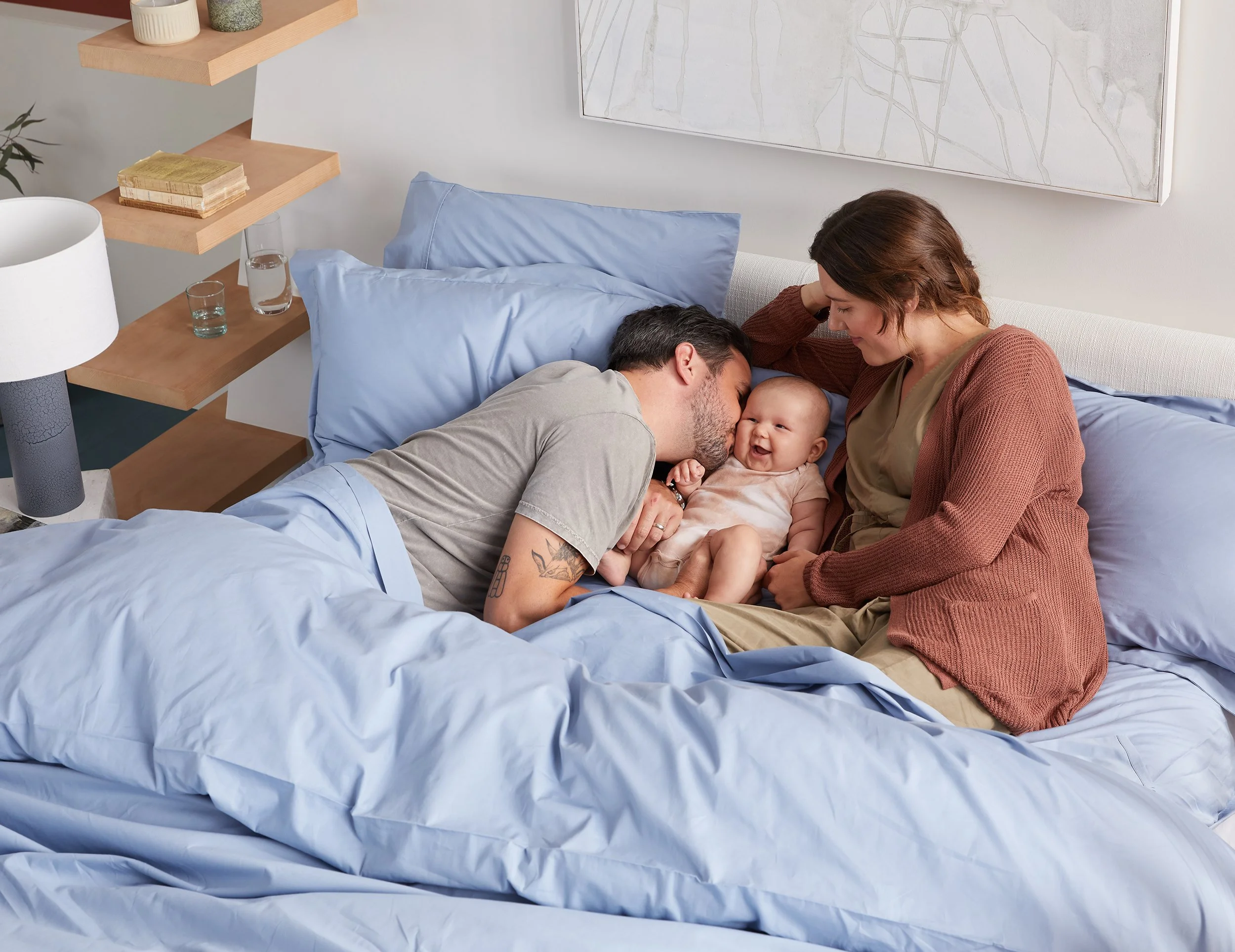 A family of three, a man, a woman, and a baby, lying on a bed in a modern bedroom. The man is kissing the baby, who is smiling, while the woman gently touches the baby's head. The bed has light blue sheets and pillows. There are wooden shelves with b