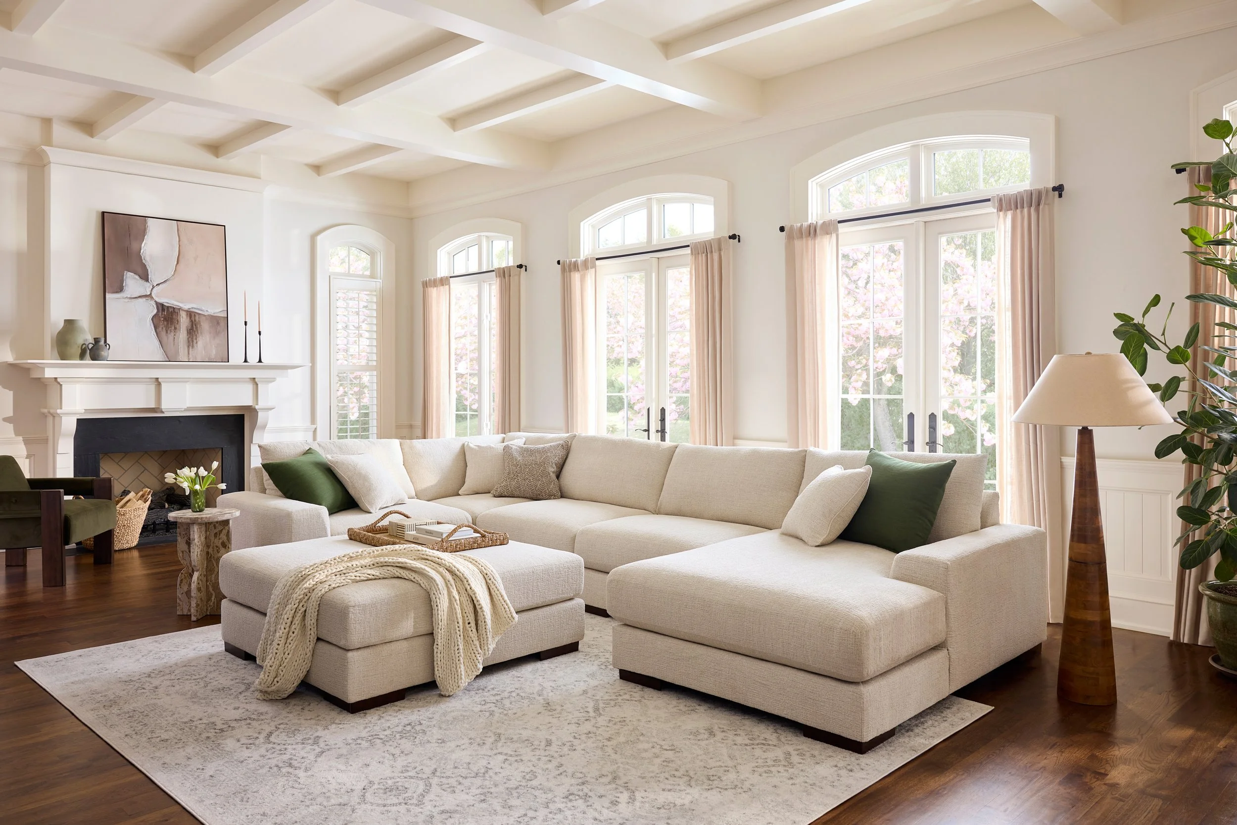 A spacious living room with a cream sectional sofa decorated with green pillows. Large windows with curtains and a view of blooming trees outside. A fireplace with decorative vases and candles on the mantel. A painting above the fireplace. A side tab