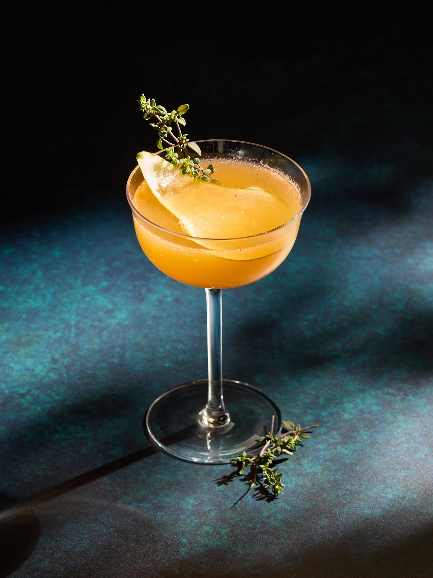A cocktail in a coupe glass garnished with a lemon wedge and fresh thyme sprigs, set against a dark background.