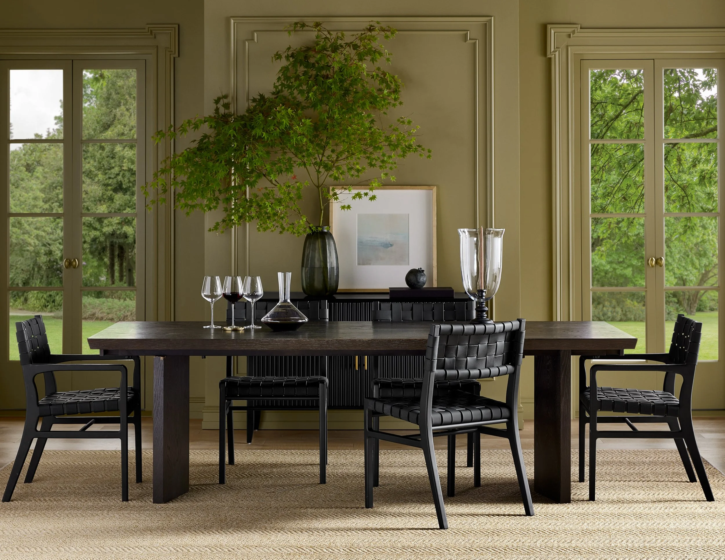 A dining room with a dark wooden table surrounded by black woven chairs. On the table are four empty wine glasses, a glass decanter filled with red wine, and tall glass candleholders. Behind the table, a large vase with green and red maple leaves is 