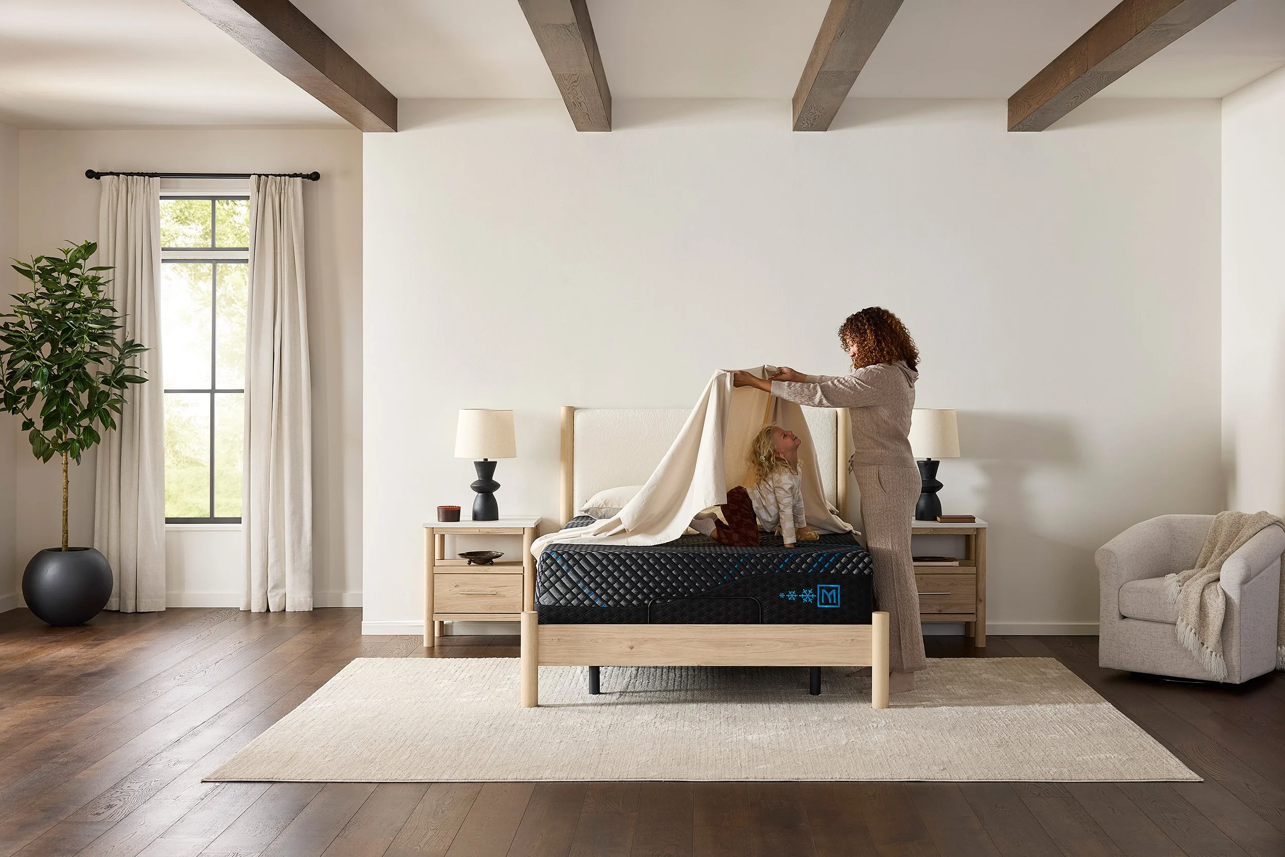 A woman and a young girl are in a bedroom. The woman appears to be making a bed or arranging a sheet over the girl, who is sitting on the bed. The bedroom has a large window with white curtains, a potted plant, two bedside tables with lamps, and wood