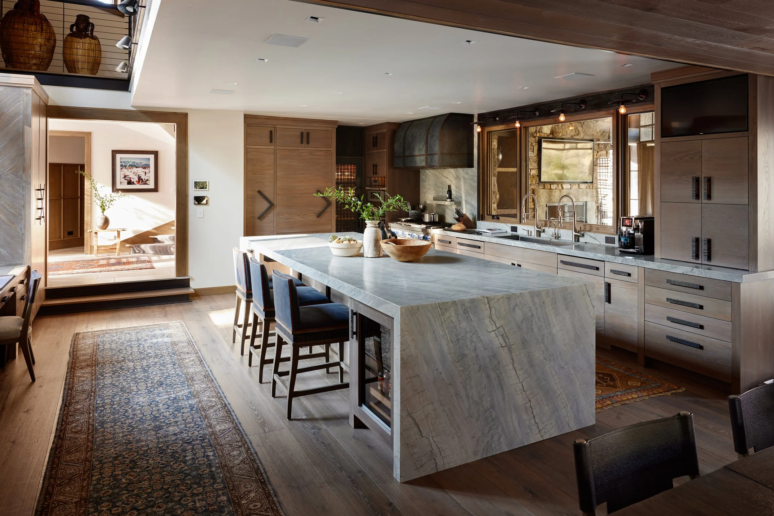 Modern kitchen with an island, cabinetry, and a stove, featuring a window with a stone wall outside, and a dining area with chairs and a rug.