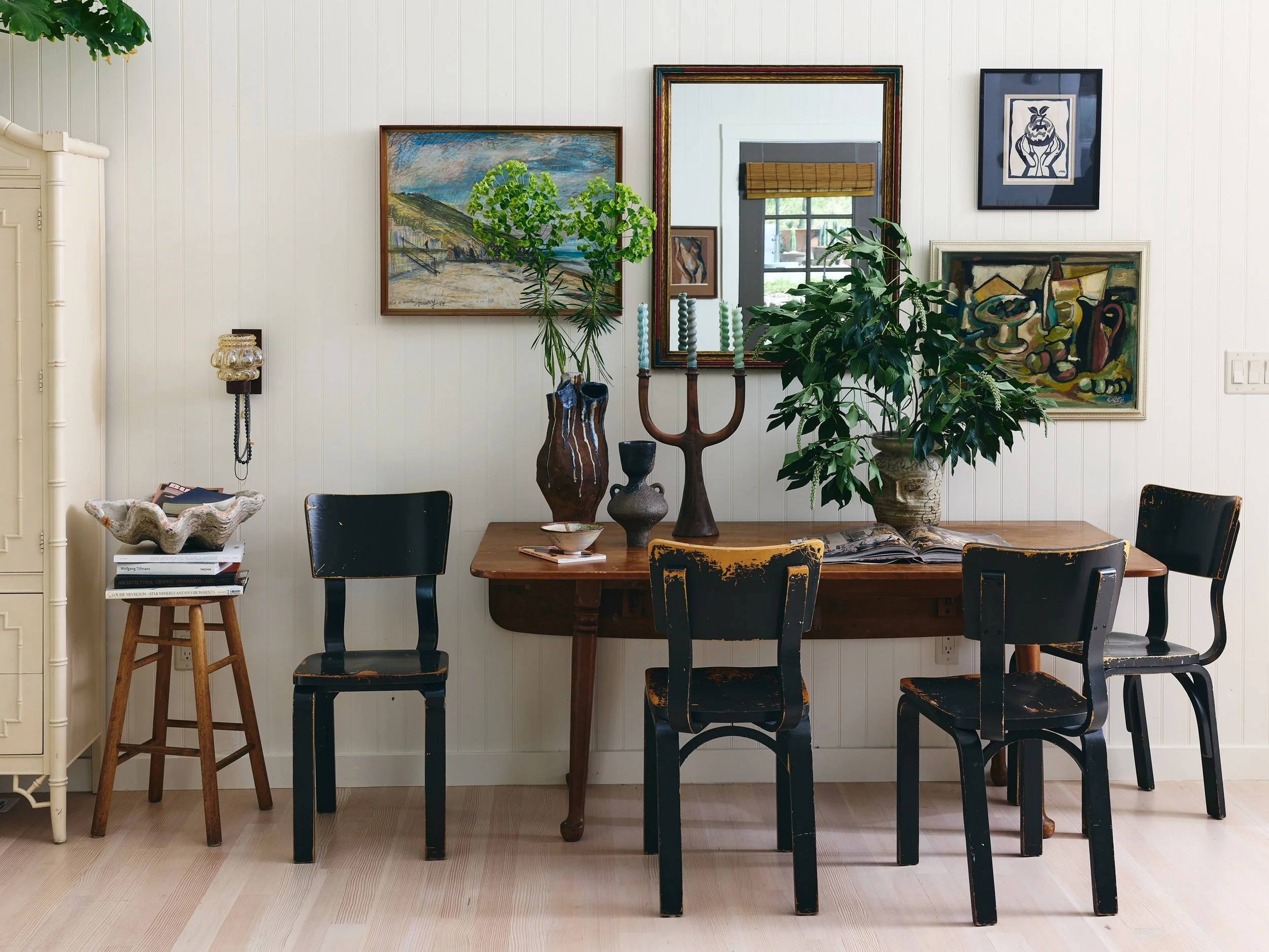 Interior of a room with white paneled walls, decorated with various framed paintings and photos. There is a long wooden table with five chairs, and decorative items such as vases, a candelabra, and a large potted plant. A side table holds books and a