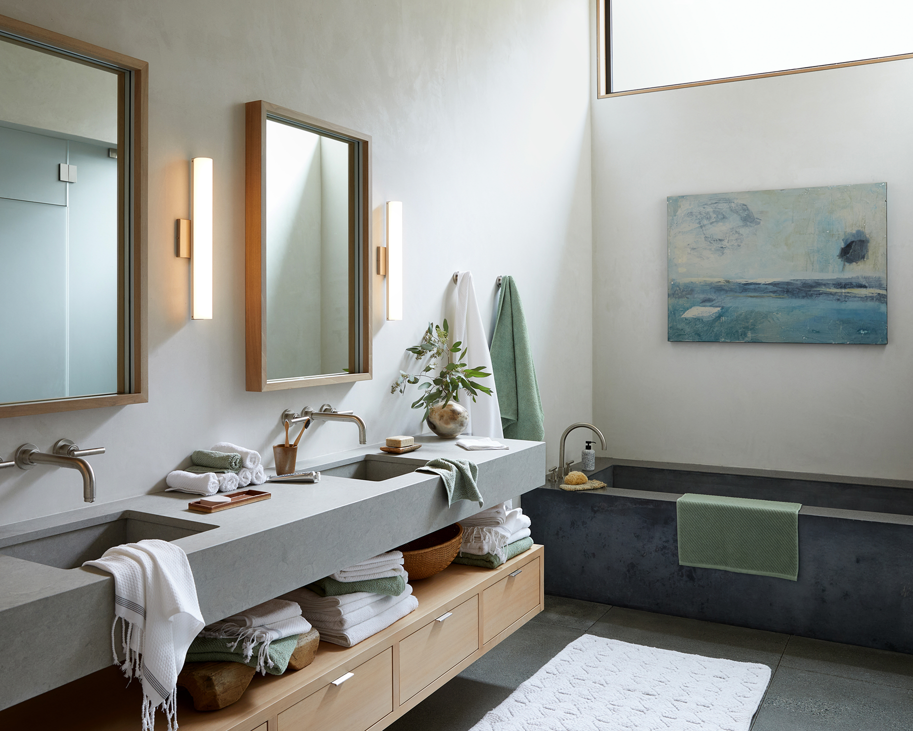 A modern bathroom with a concrete vanity, dual sinks, and a large mirror. The space features wooden accents, green and white towels, plants, and wall-mounted lighting. There is a soaking tub with a towel hanging over it and a white textured rug on th