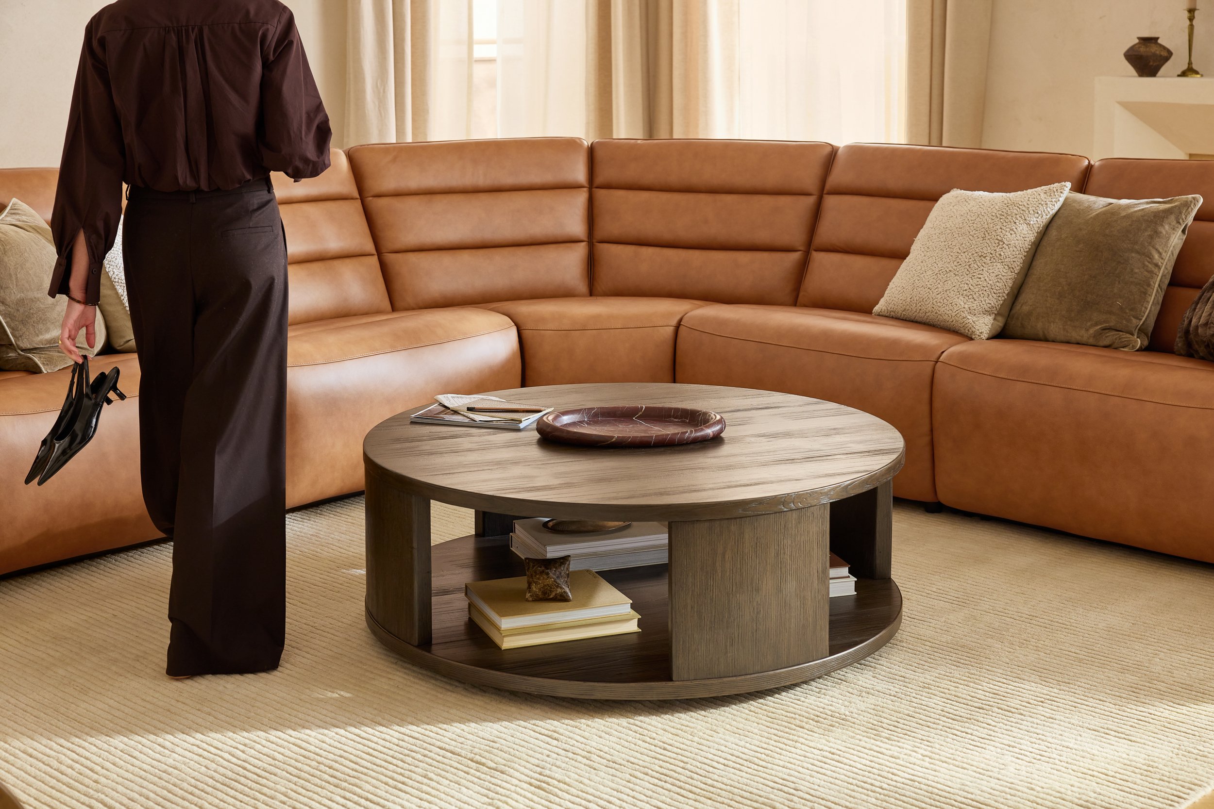 A person standing in a living room with a brown leather sectional sofa, a round wooden coffee table with books and decorative items, and beige curtains covering windows.