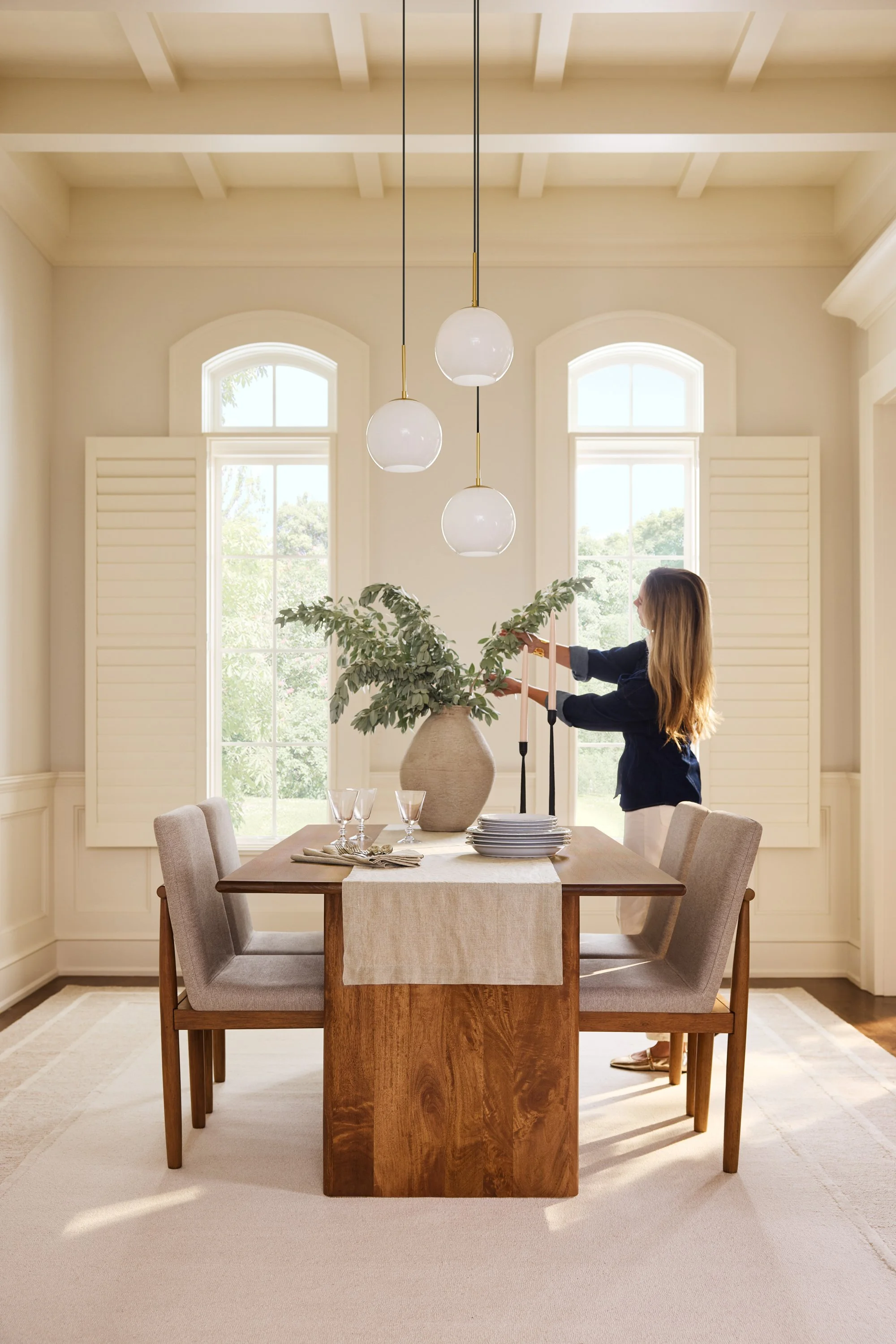 A woman arranging a plant on a dining table in a bright room with large windows, hanging pendant lights, and beige chairs.