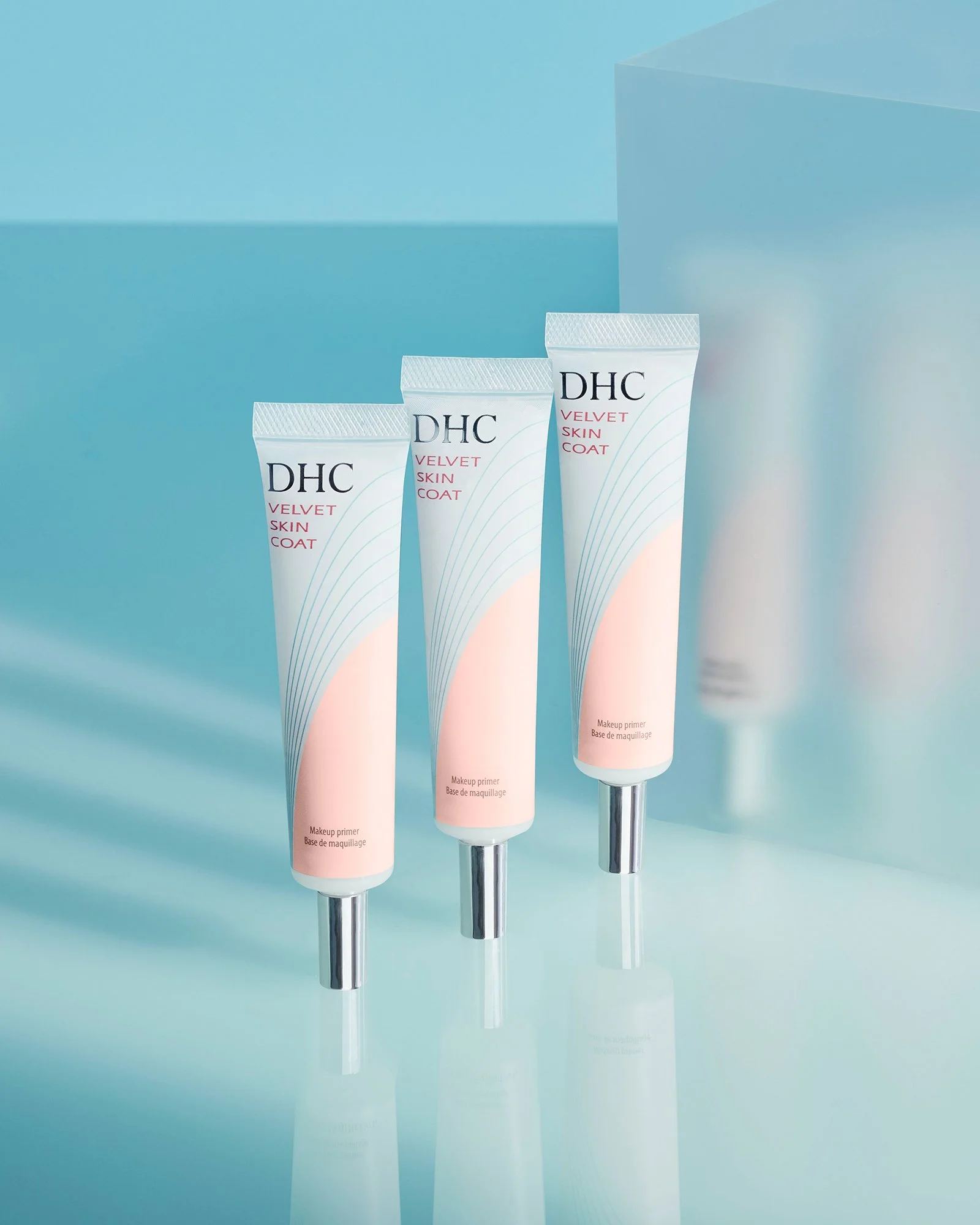 Three tubes of DHC Velvet Skin Coat makeup primer on a surface with a light blue background.