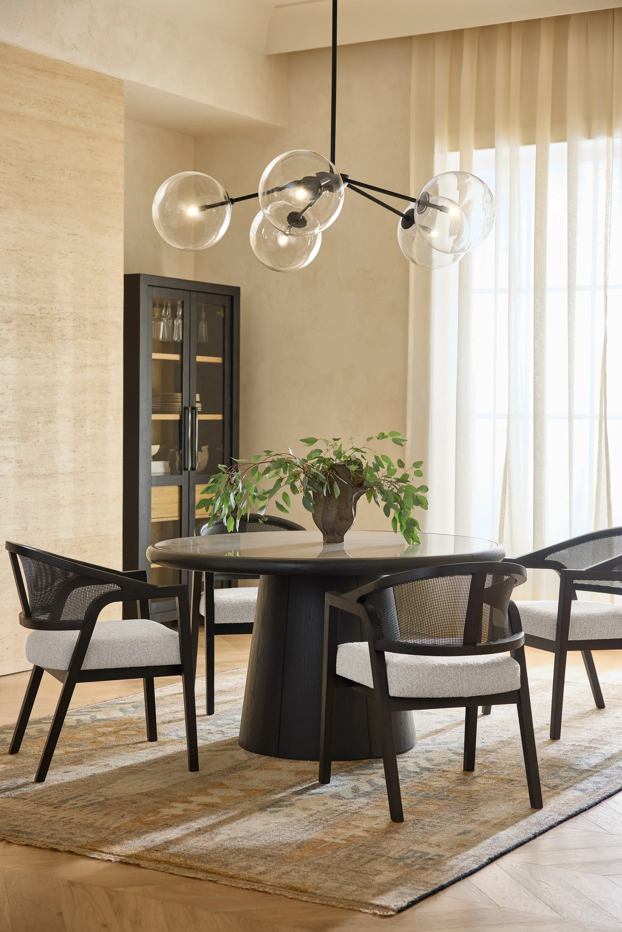 Modern dining room with a round table, four black chairs with white cushions, a dark wood cabinet with glass door, large window with sheer curtains, a vase with green leaves on the table, and a contemporary chandelier with clear globes hanging from b