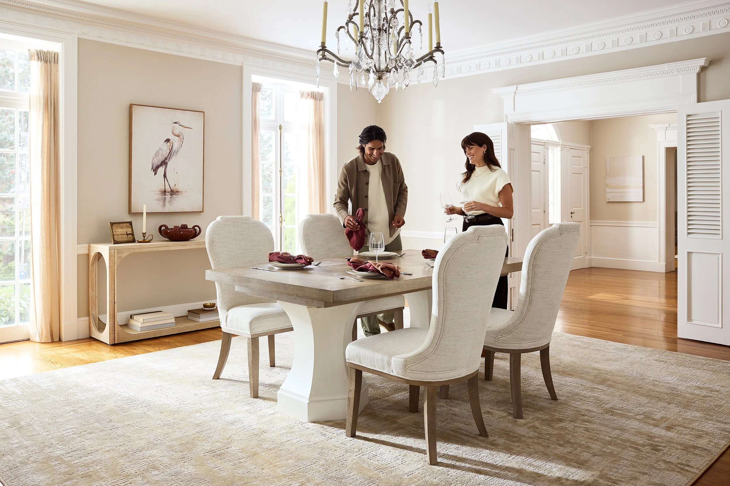 A dining room with a wooden table set with wine glasses and napkins, surrounded by upholstered chairs, in a bright room with large windows, a chandelier, and light-colored walls with artwork.