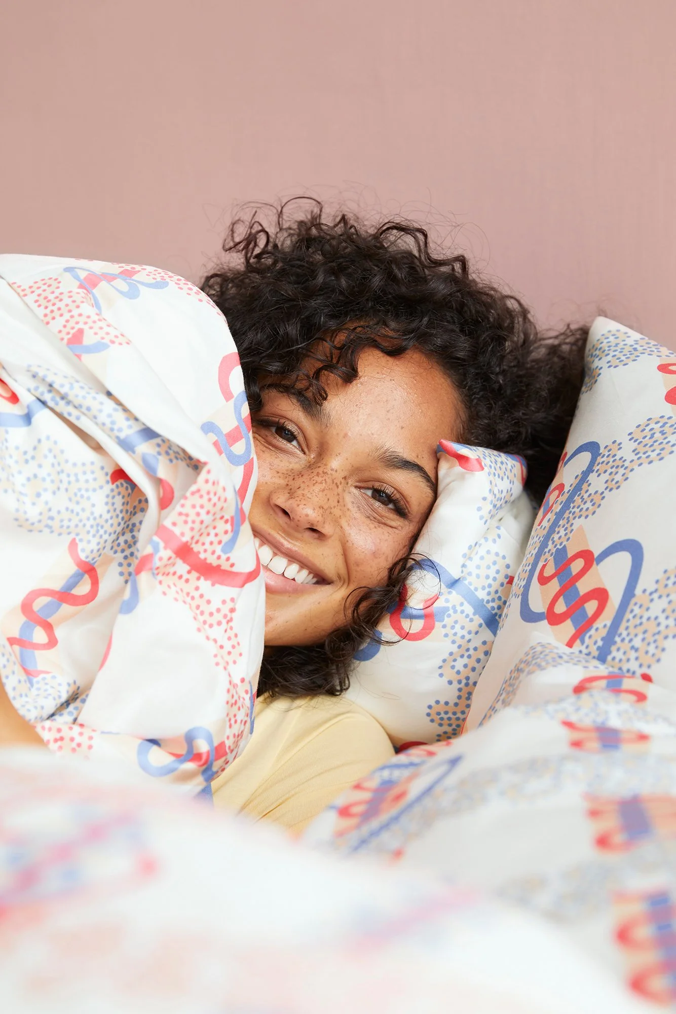 Person with curly hair smiling and lying on a bed with patterned sheets and pillows.