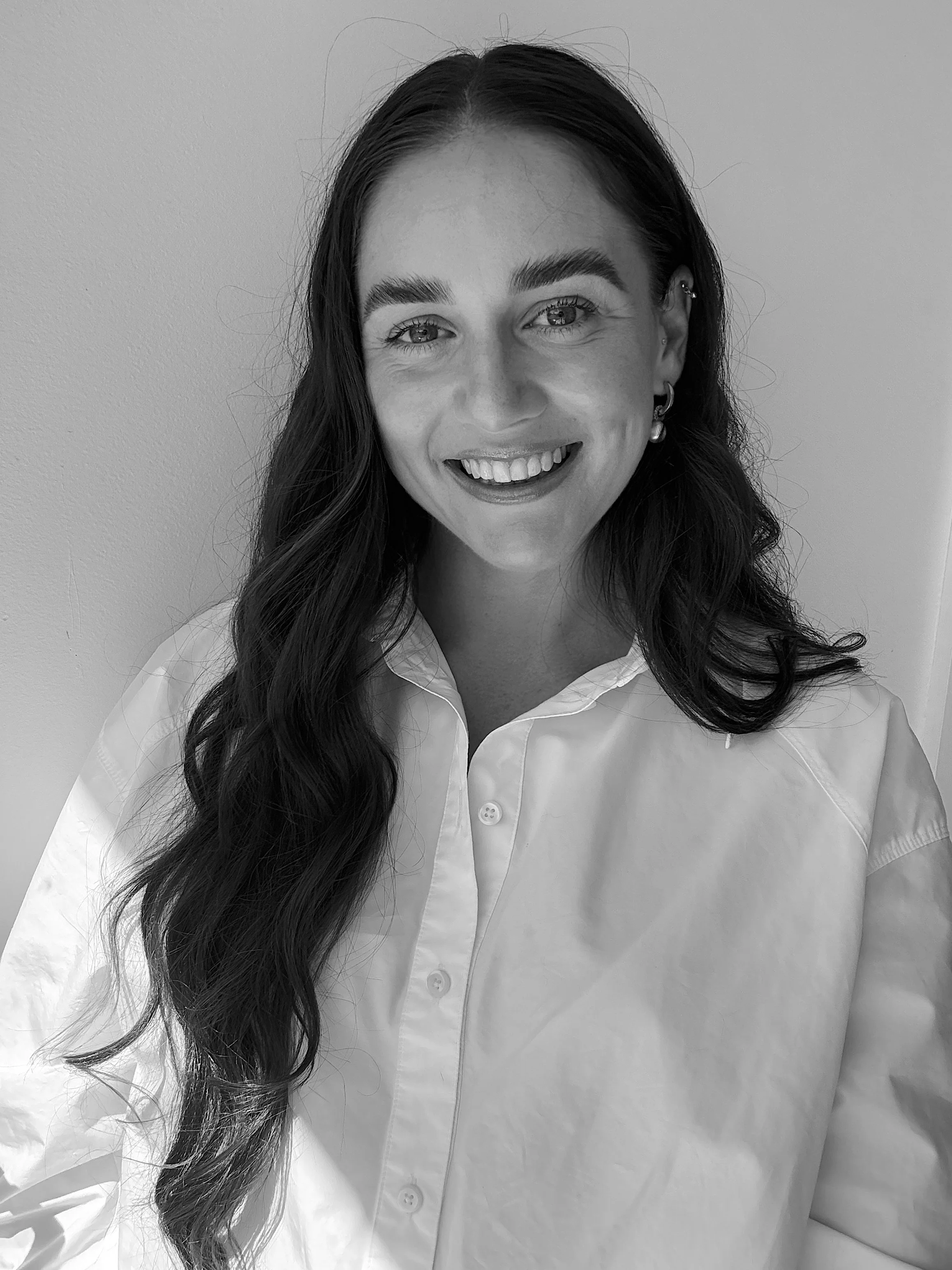 Black and white portrait of a woman with long wavy hair wearing a white button-up shirt, smiling, standing against a plain light-colored wall.
